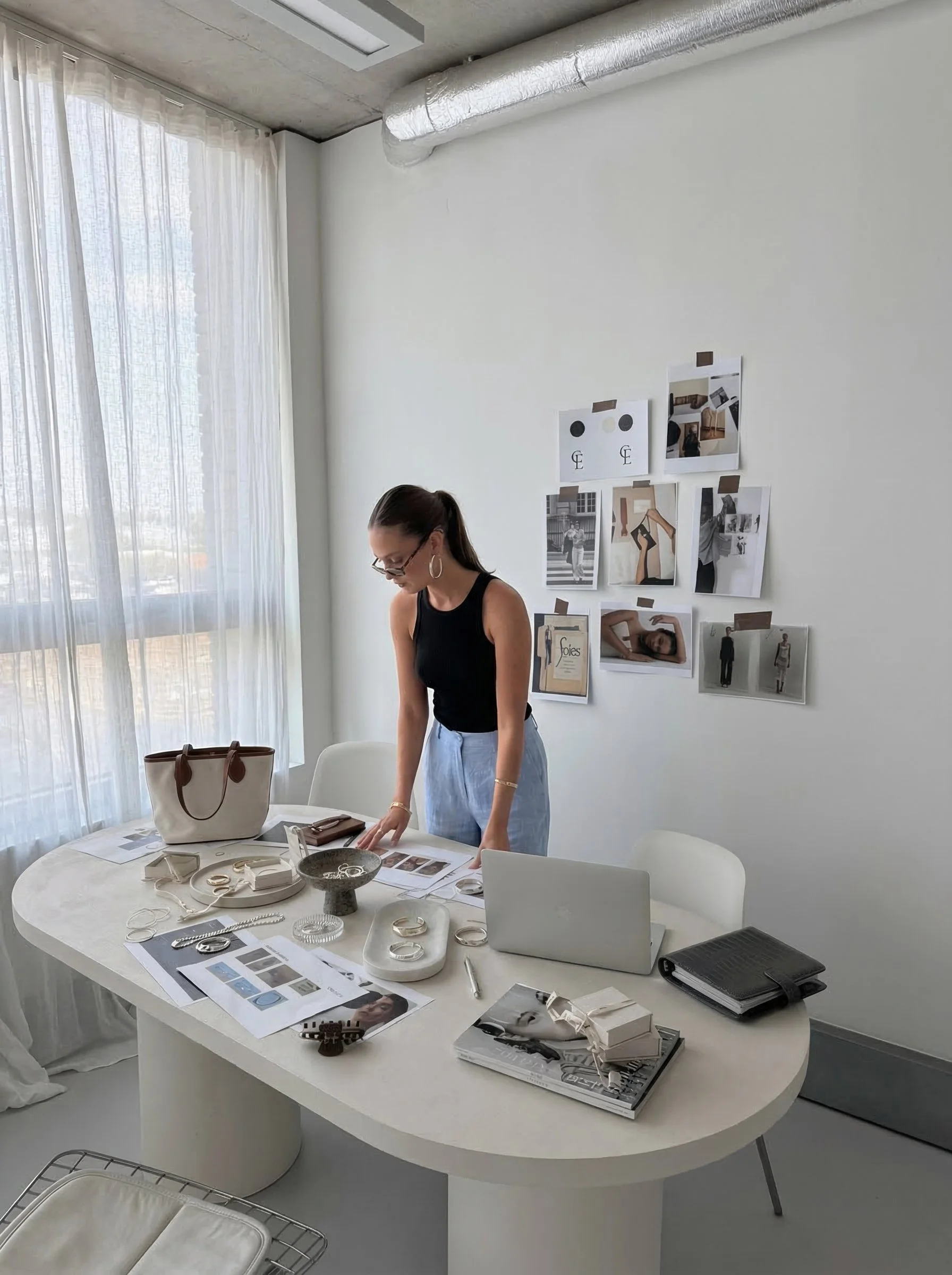 A woman standing at a white oval table covered with jewelry, papers, and a laptop in a bright room with natural light coming through sheer white curtains. Behind her, a white wall has a collage of photographs and images taped to it.