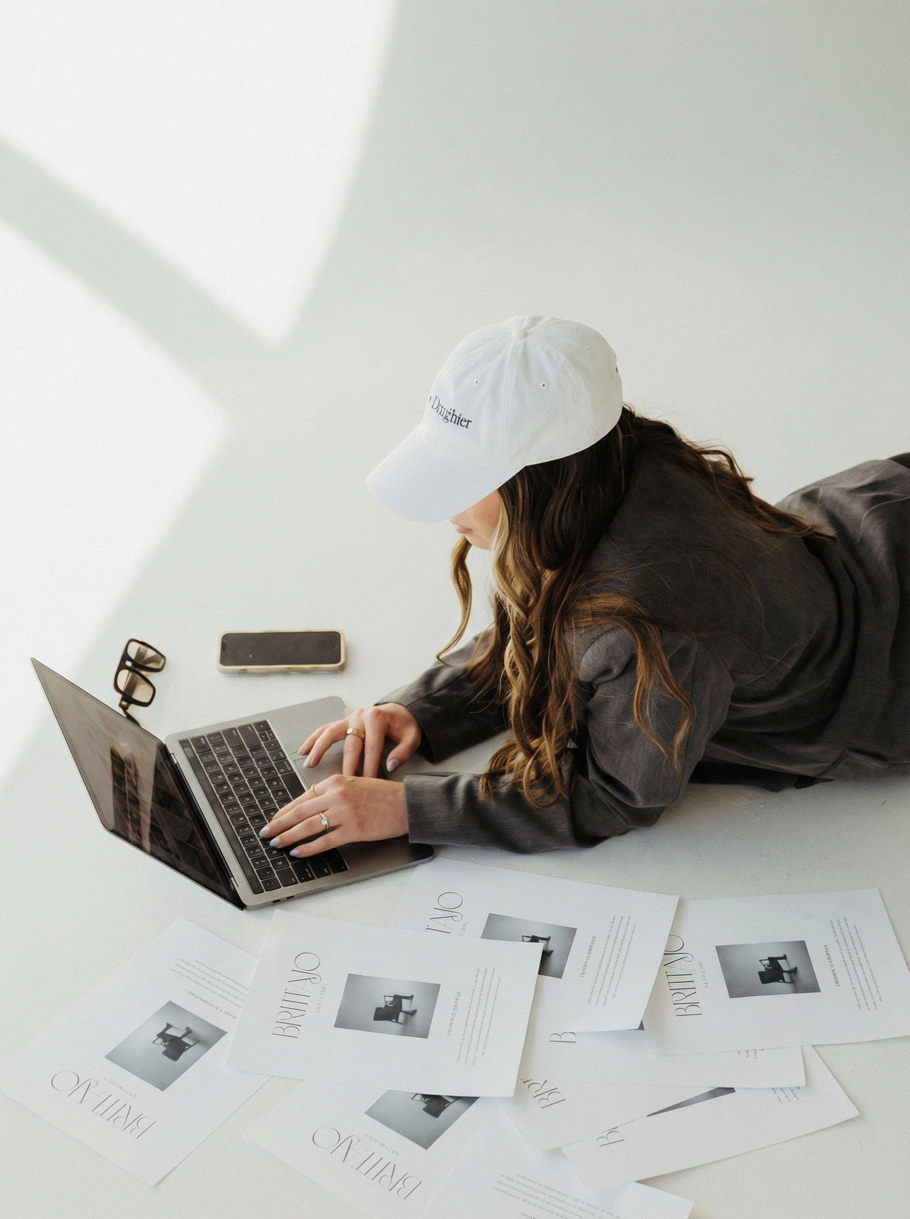 A woman lying on the floor working on a laptop, with papers and a smartphone surrounding her, wearing a white cap and a dark jacket.