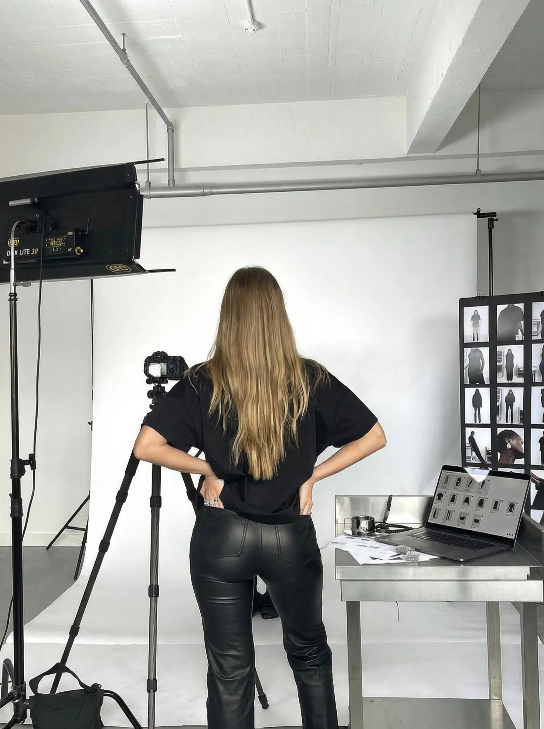 A woman with long blonde hair stands in a photography studio, facing away from the camera. She is wearing a black T-shirt and black leather pants, with her hands on her hips. There is a camera on a tripod facing her, a laptop opened on a table, and a large screen displaying multiple images of people in the background.