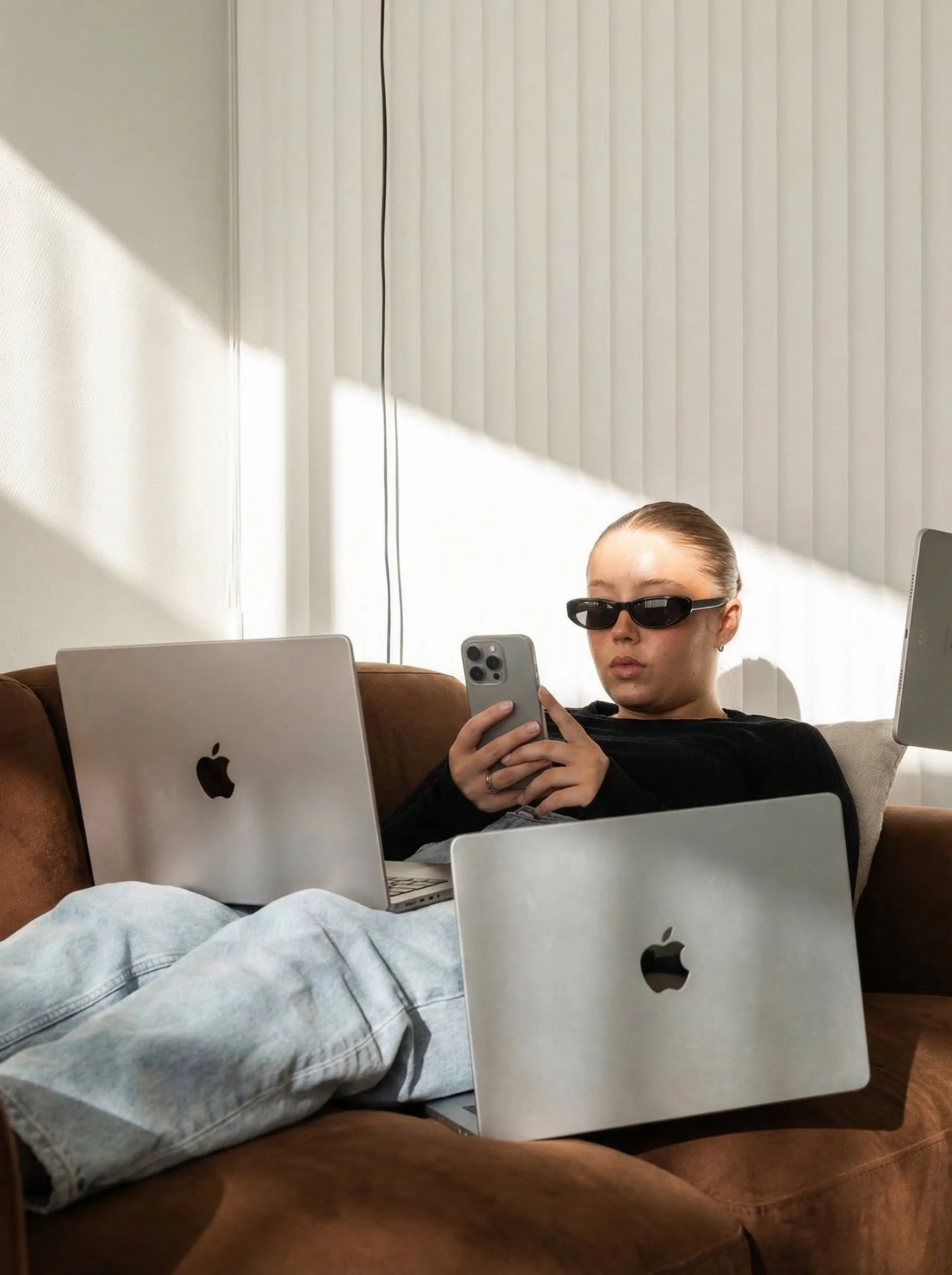 A woman wearing sunglasses is sitting on a brown couch with multiple Apple laptops around her, holding a smartphone and looking at it. She is dressed in casual clothing with light-colored jeans and a black top.