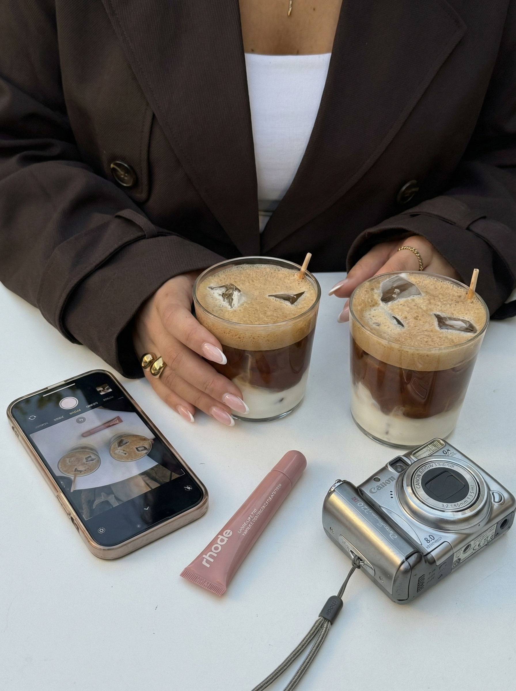 Person wearing a brown blazer holding two iced coffee drinks with layered milk and coffee, on a white table alongside a smartphone showing the same drinks, a pink Rhode balm, and a silver Canon camera.