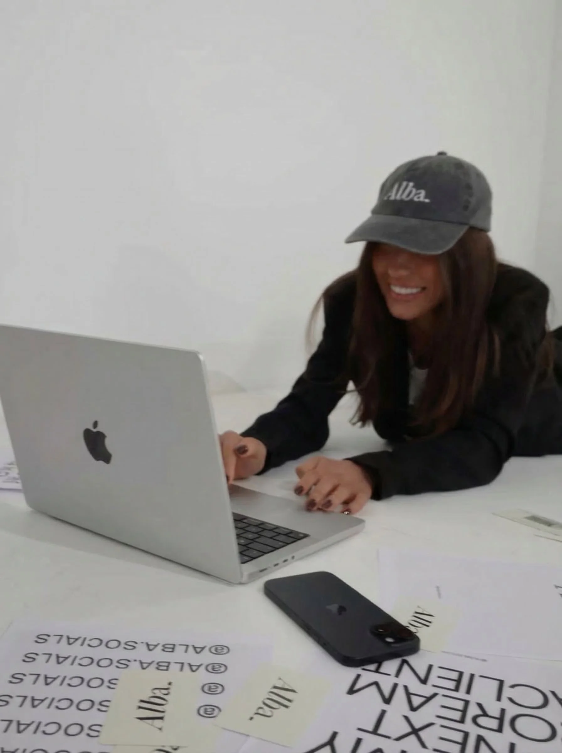 Woman lying on a table with a laptop and papers, smiling, wearing a gray cap and black jacket.