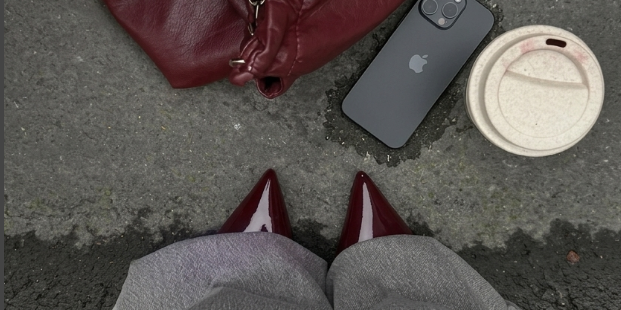 Person standing on sidewalk wearing gray pants and red high heels, with a red purse, an iPhone, and coffee cup on the ground.