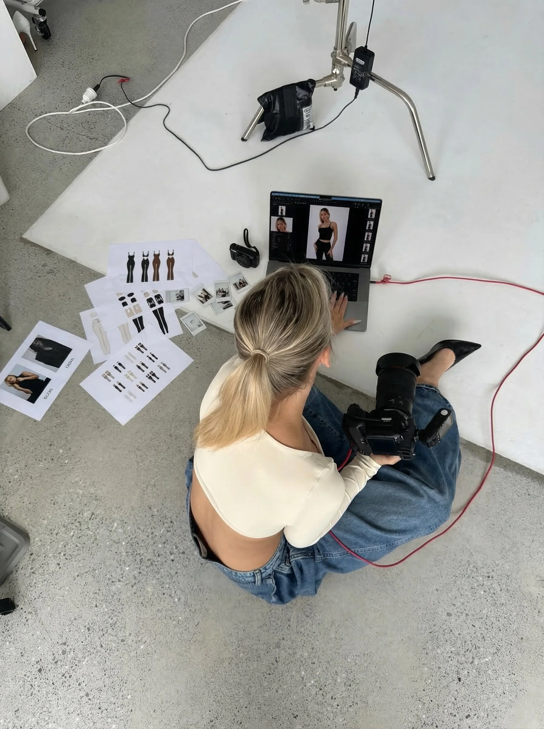 A woman sitting on the floor taking photos of a model wearing various dresses using a camera and laptop in a studio with clothing images and photos spread on the floor.