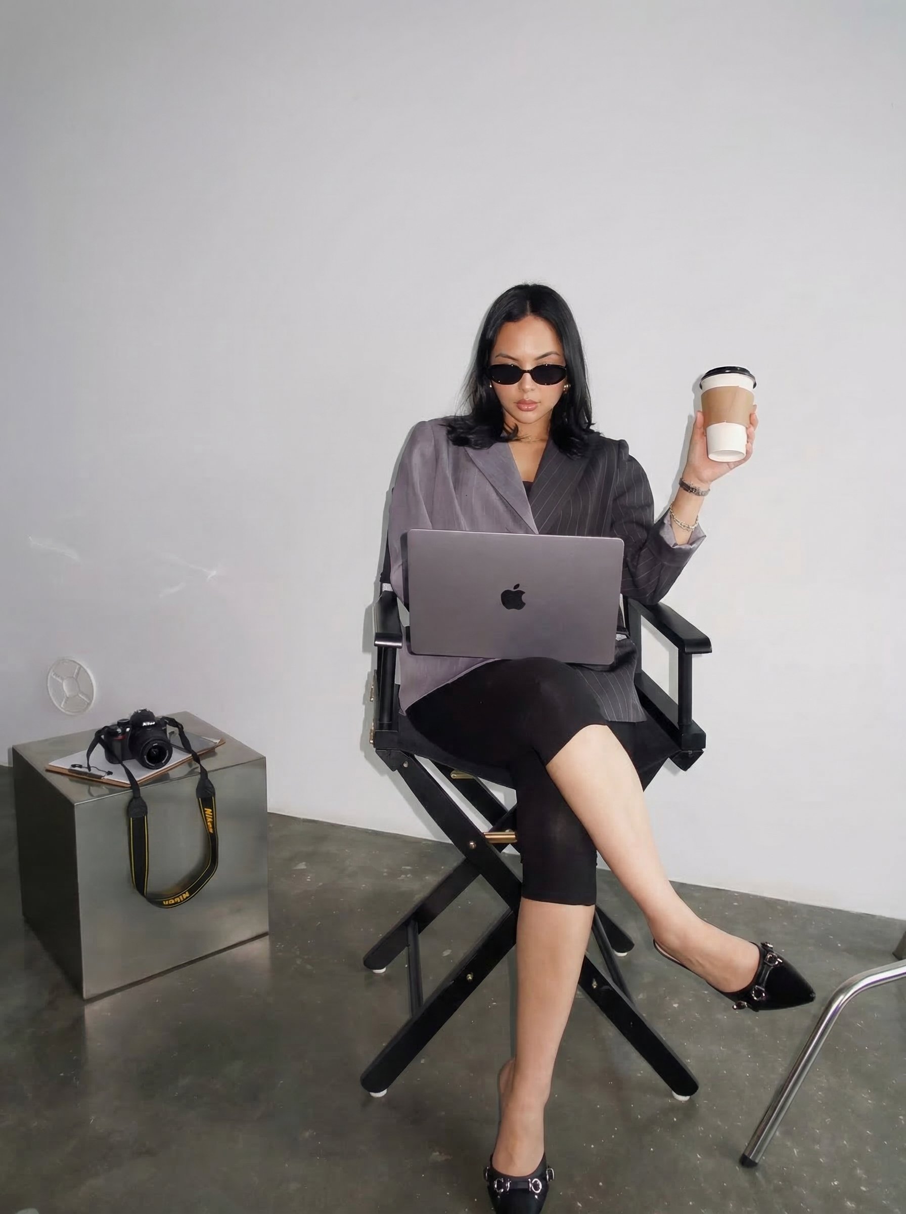 A woman sitting on a director's chair working on a laptop, holding a coffee cup, wearing sunglasses, a pinstripe blazer, black skirt and heels, with a camera on a nearby table, against a plain white wall.