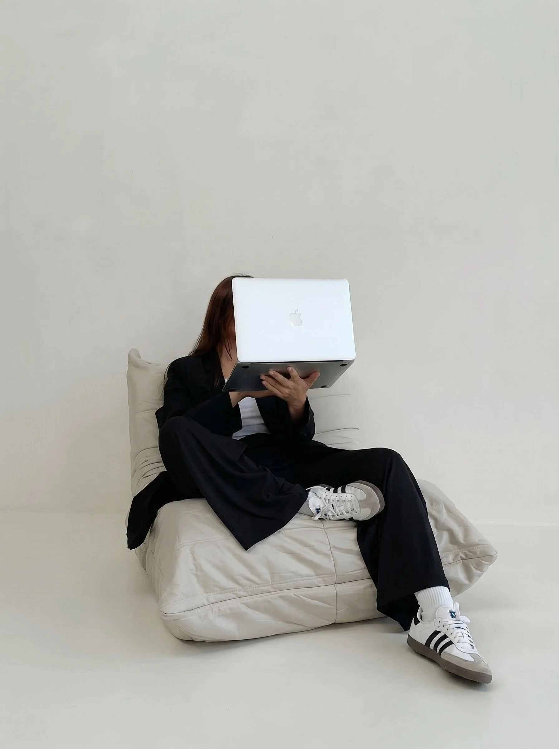 Person sitting on a bed, holding a laptop in front of their face, with a plain white background.