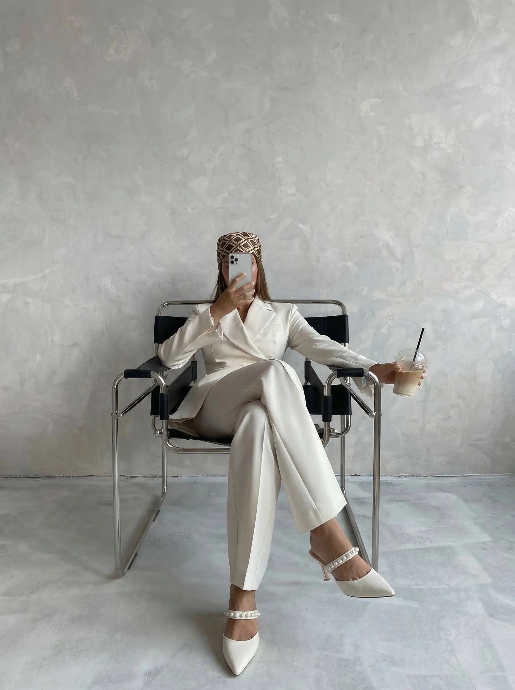 A woman sitting on a modern black and silver chair, wearing a cream-colored suit with pearl-embellished high heels, holding a cold coffee with a straw in one hand, and taking a selfie with her phone while wearing a patterned headscarf, against a plain, light gray wall.