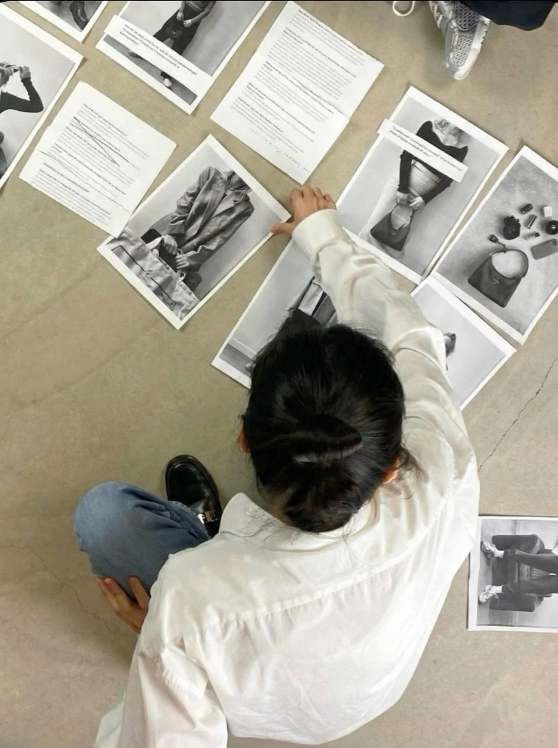 A person with dark hair tied up, wearing a white shirt and blue jeans, sitting on the floor, pointing at black-and-white photographs and printed documents laid out around them. The images include fashion models, objects, and text.