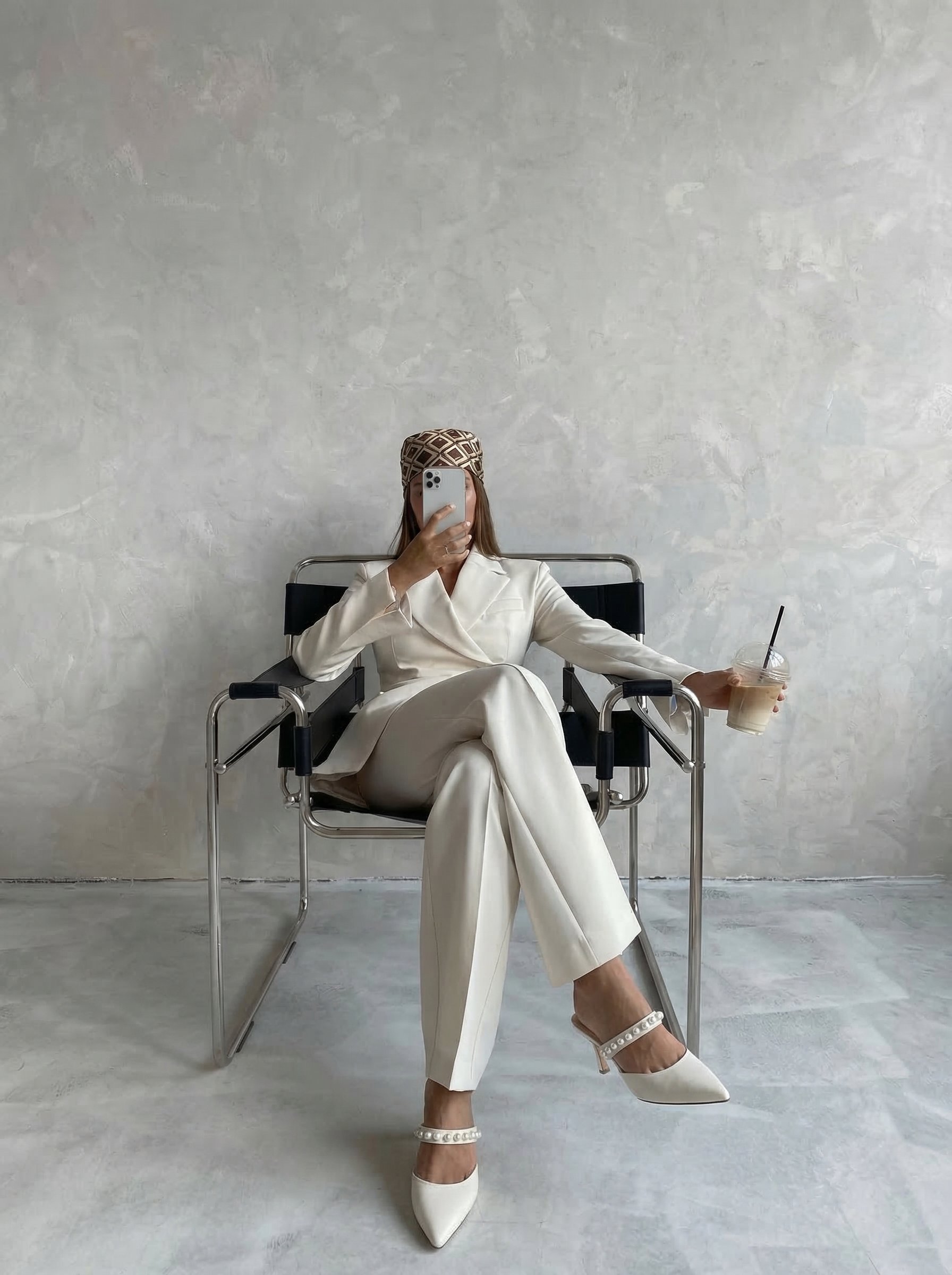 Woman in a cream-colored suit and heels sitting on a black chair with metal frame, taking a selfie with her phone, holding a cup with a straw, seated against a plain light-colored textured wall.