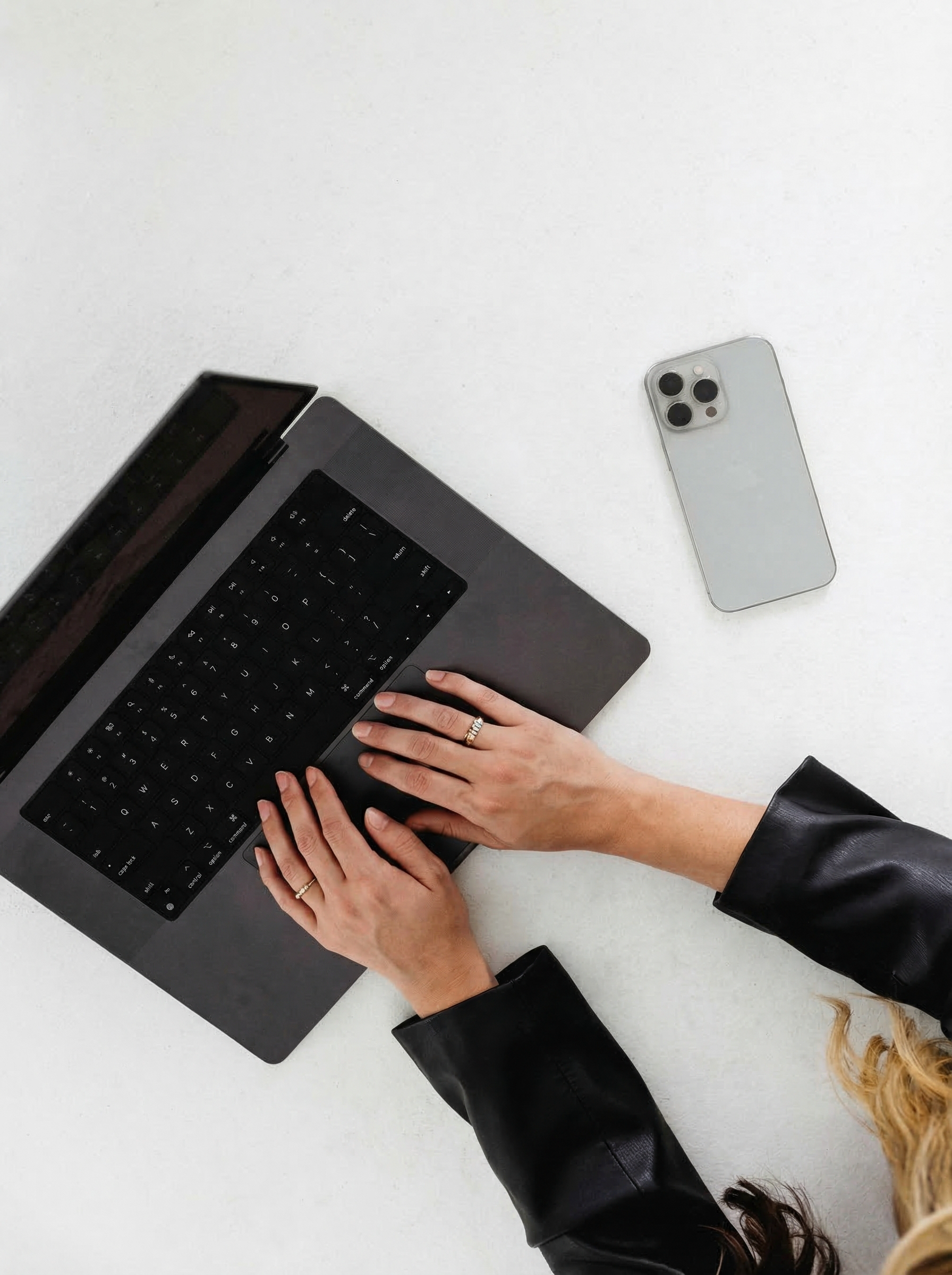 Person typing on a laptop keyboard with a smartphone on a white surface nearby.