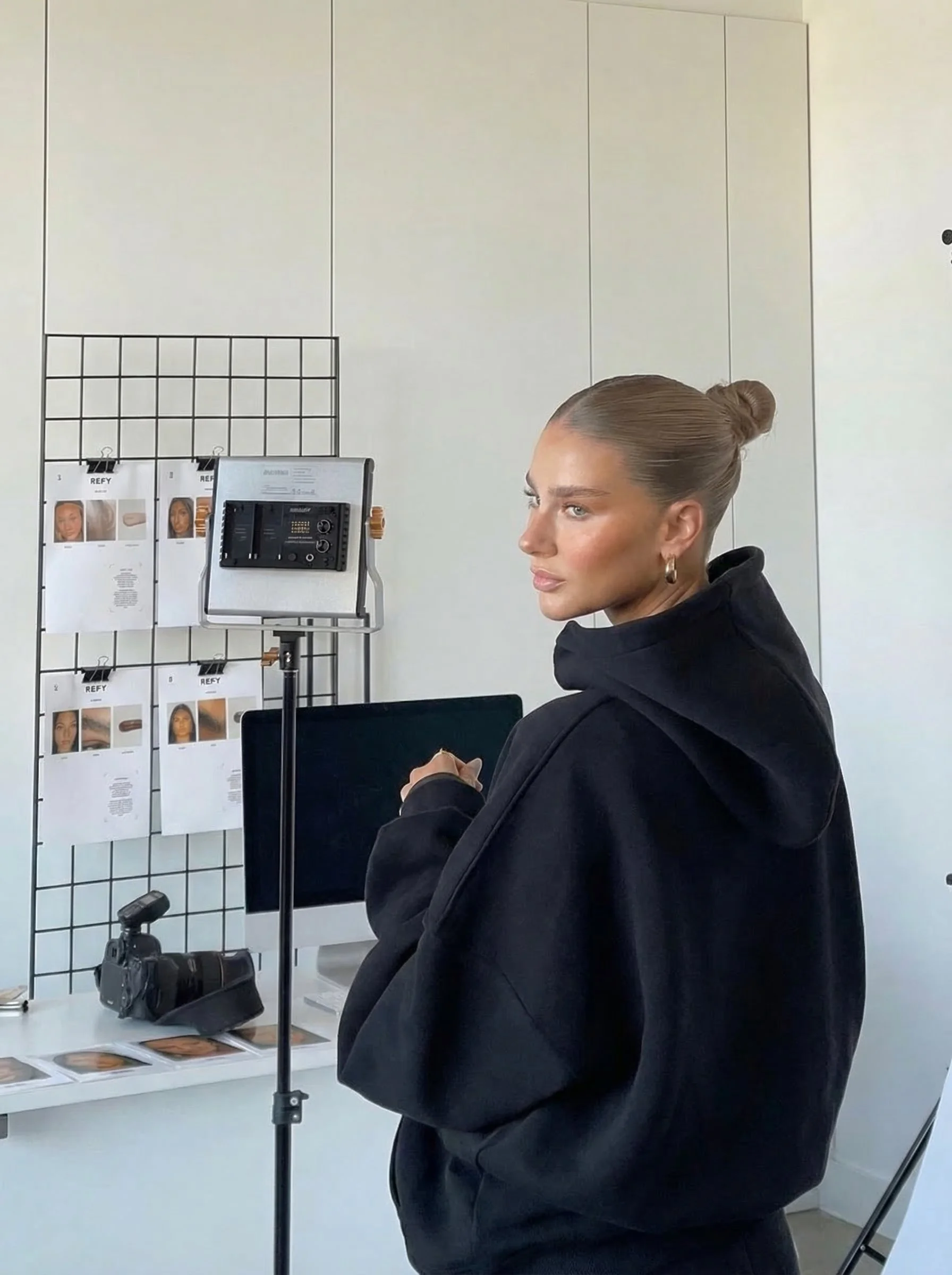 A woman with blonde hair styled in a bun is looking to her left, wearing a black hoodie, in a room with a white wall behind her and a display of papers and a camera on the table.