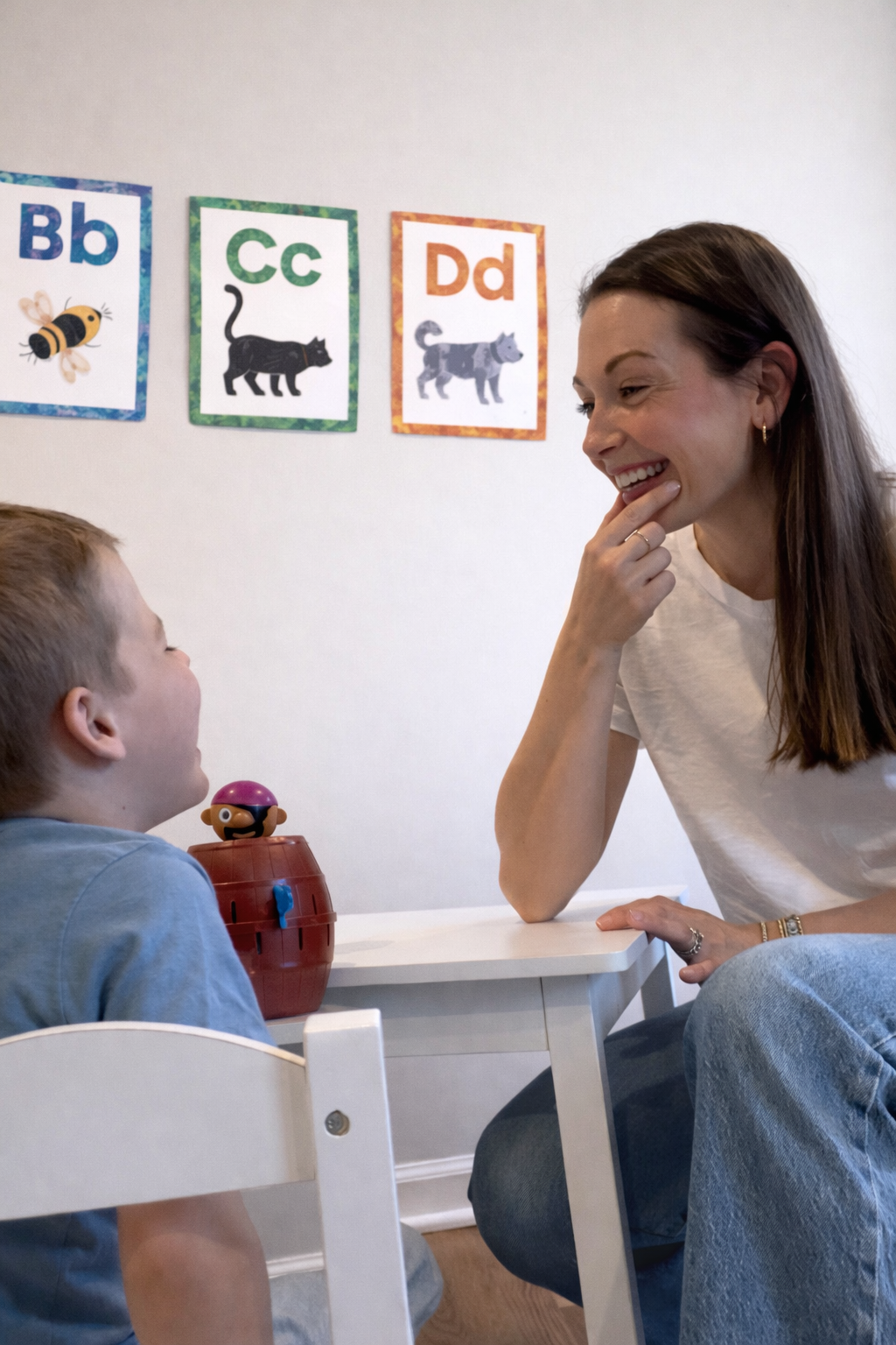 A young boy and an adult woman are sitting at a small table in a classroom, smiling and engaging in conversation. Behind them on the wall are colorful alphabet posters with pictures of animals and letters. Speech therapy Ambler, PA.