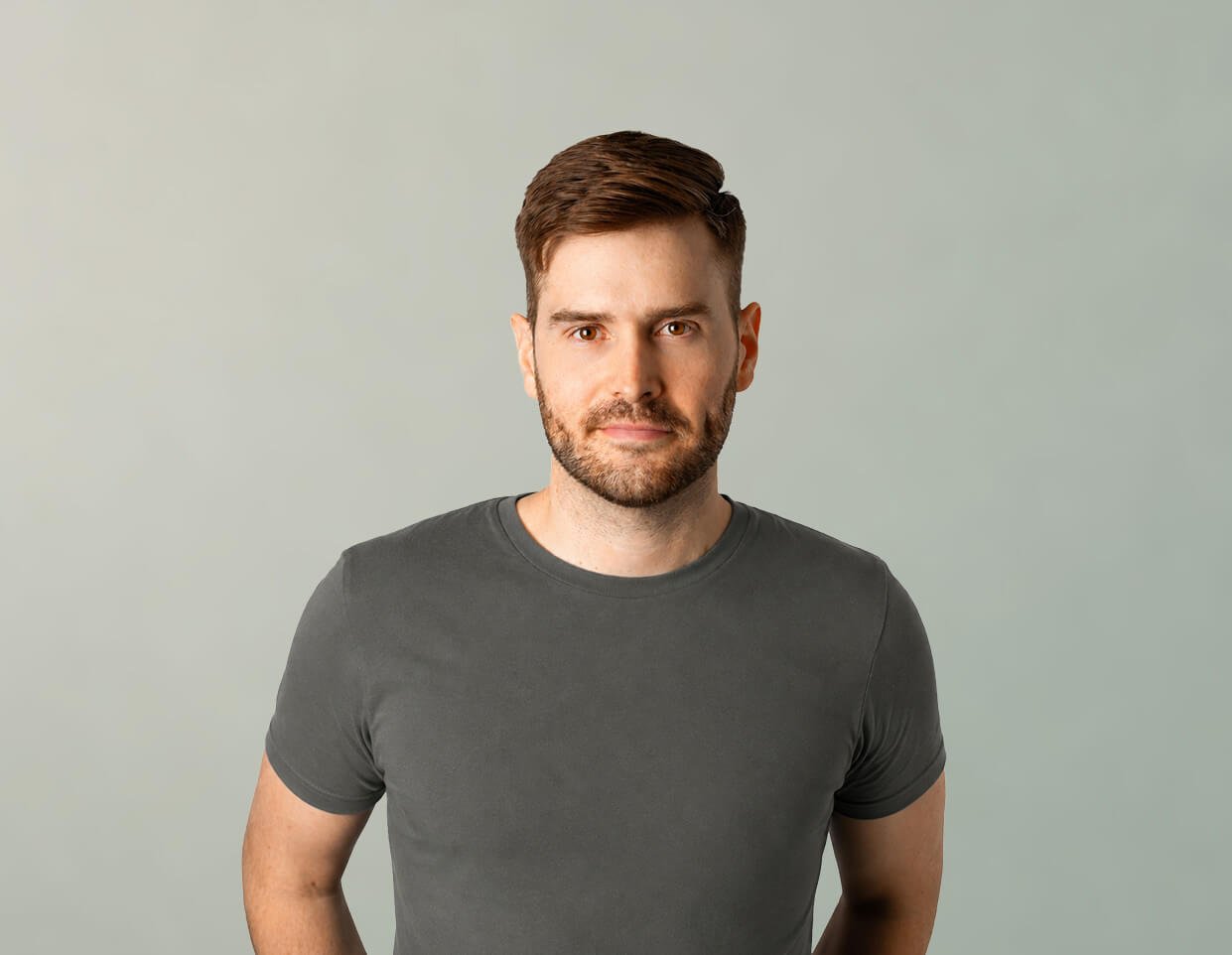 A man with brown hair, beard, and mustache wearing a dark gray t-shirt, looking at the camera with a neutral expression against a light green background.