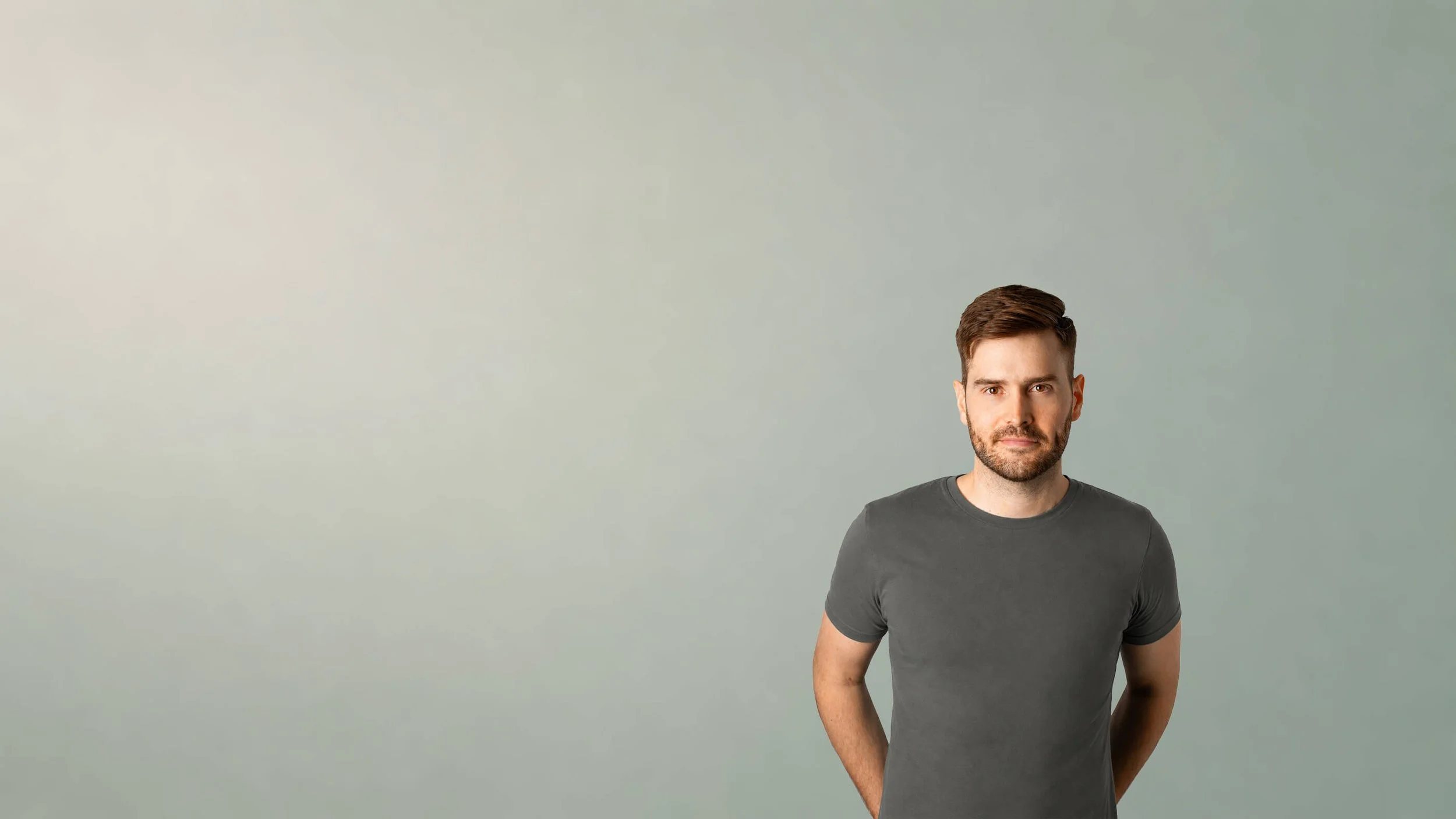 A man with short brown hair and a beard standing against a light green background, wearing a dark gray T-shirt.