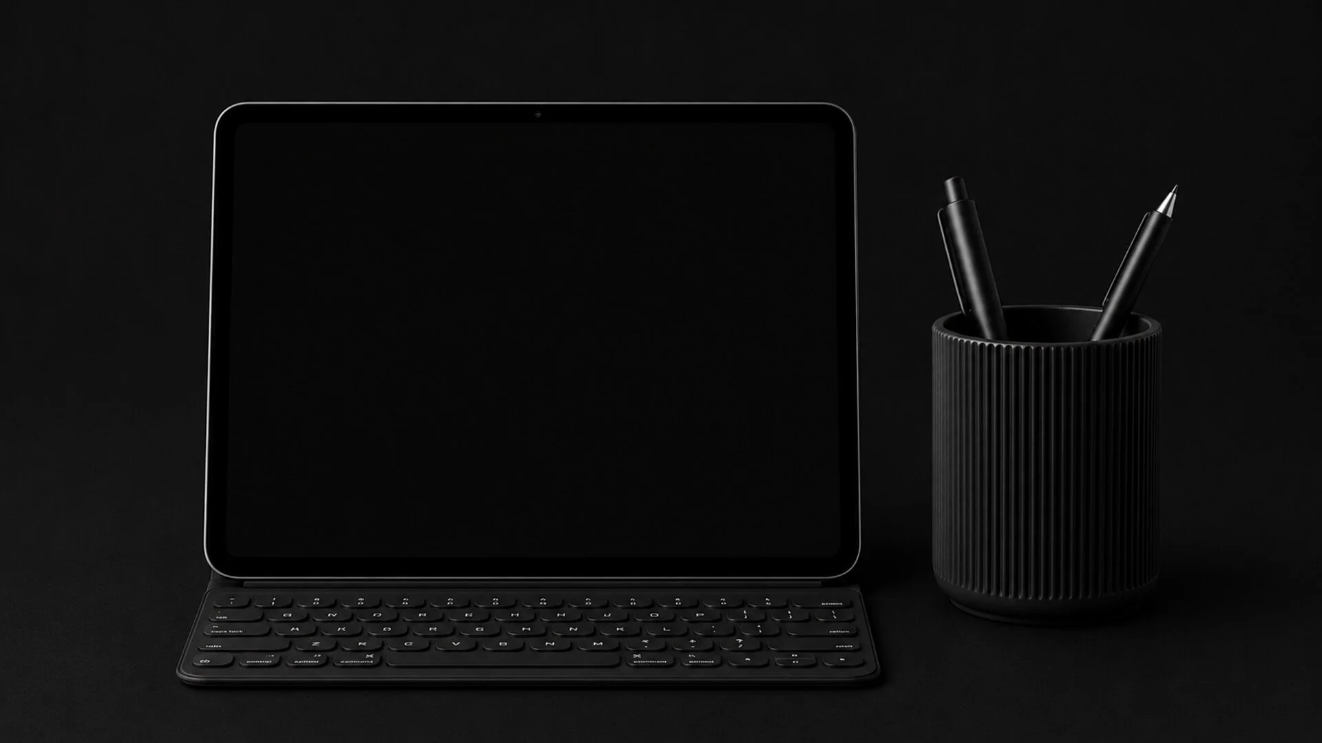 Black tablet with detachable keyboard next to a black ribbed pen holder with two black pens on a black background.