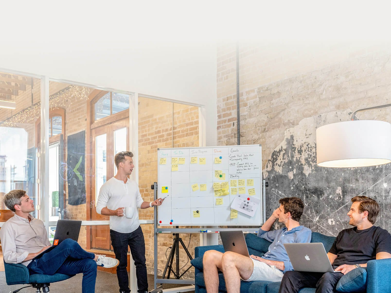 A group of five men in a modern office space during a meeting. One man stands in front of a whiteboard filled with sticky notes and notes, holding a cup and pointing. The others are seated, some with laptops, engaged and listening. The room has brick walls, large windows, and a large hanging lamp.
