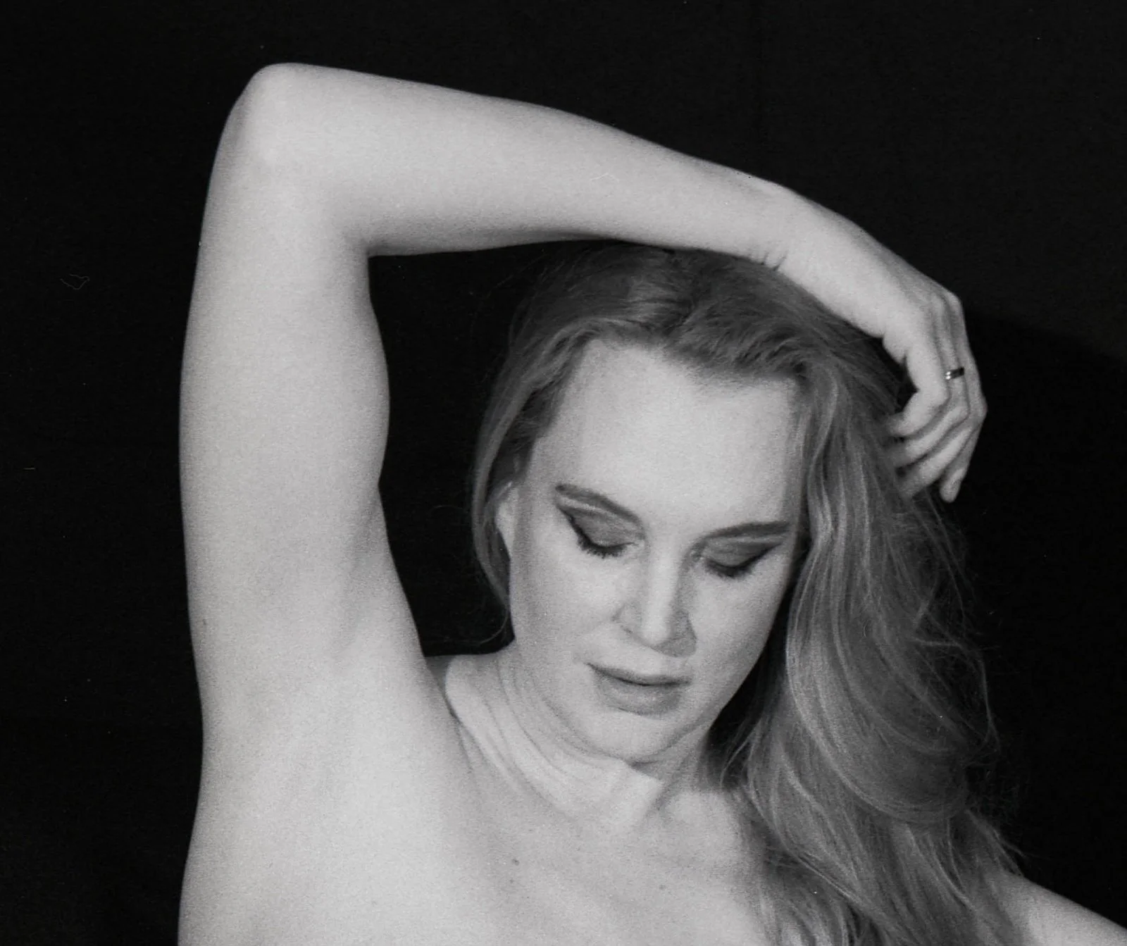 Black-and-white analogue portrait of Ariel Anderssen, bare-shouldered with one arm raised above her head and her gaze lowered against a dark background.