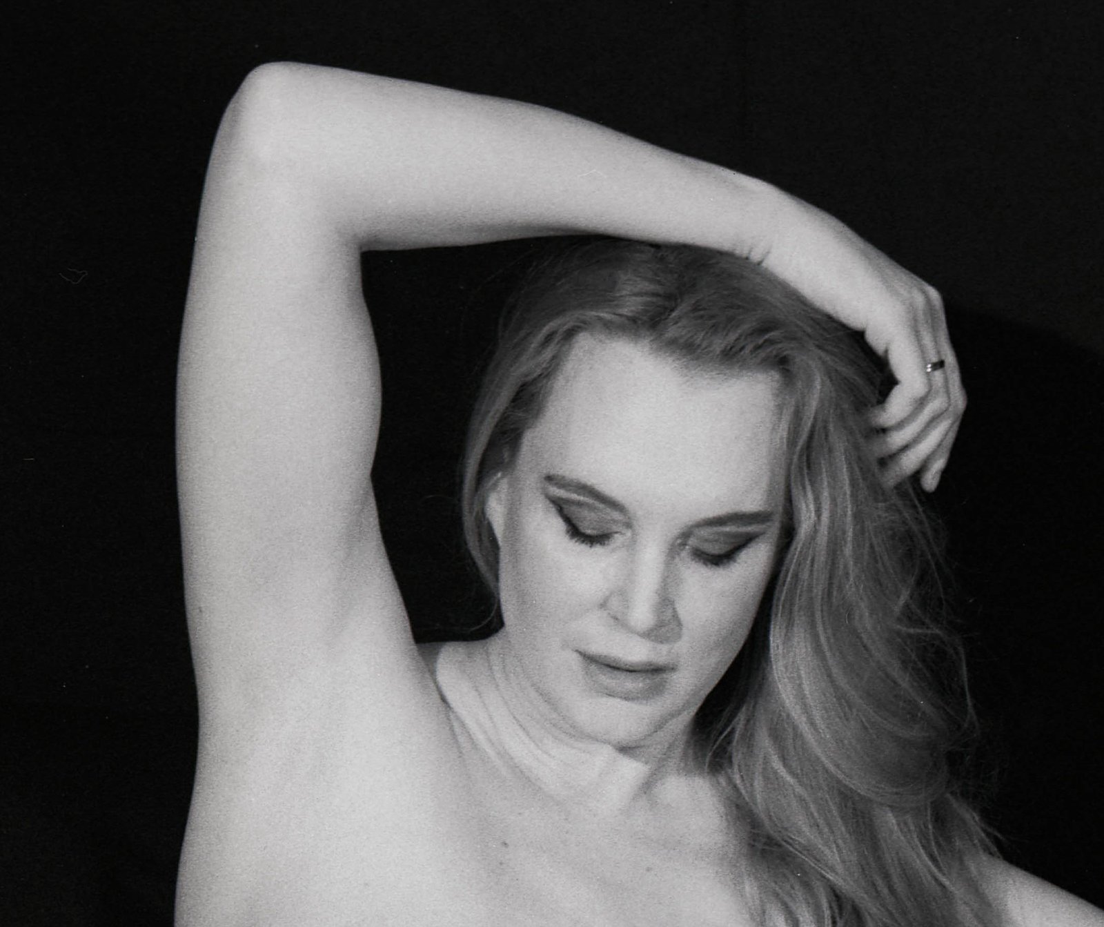 Black-and-white analogue portrait of Ariel Anderssen, bare-shouldered with one arm raised above her head and her gaze lowered against a dark background.