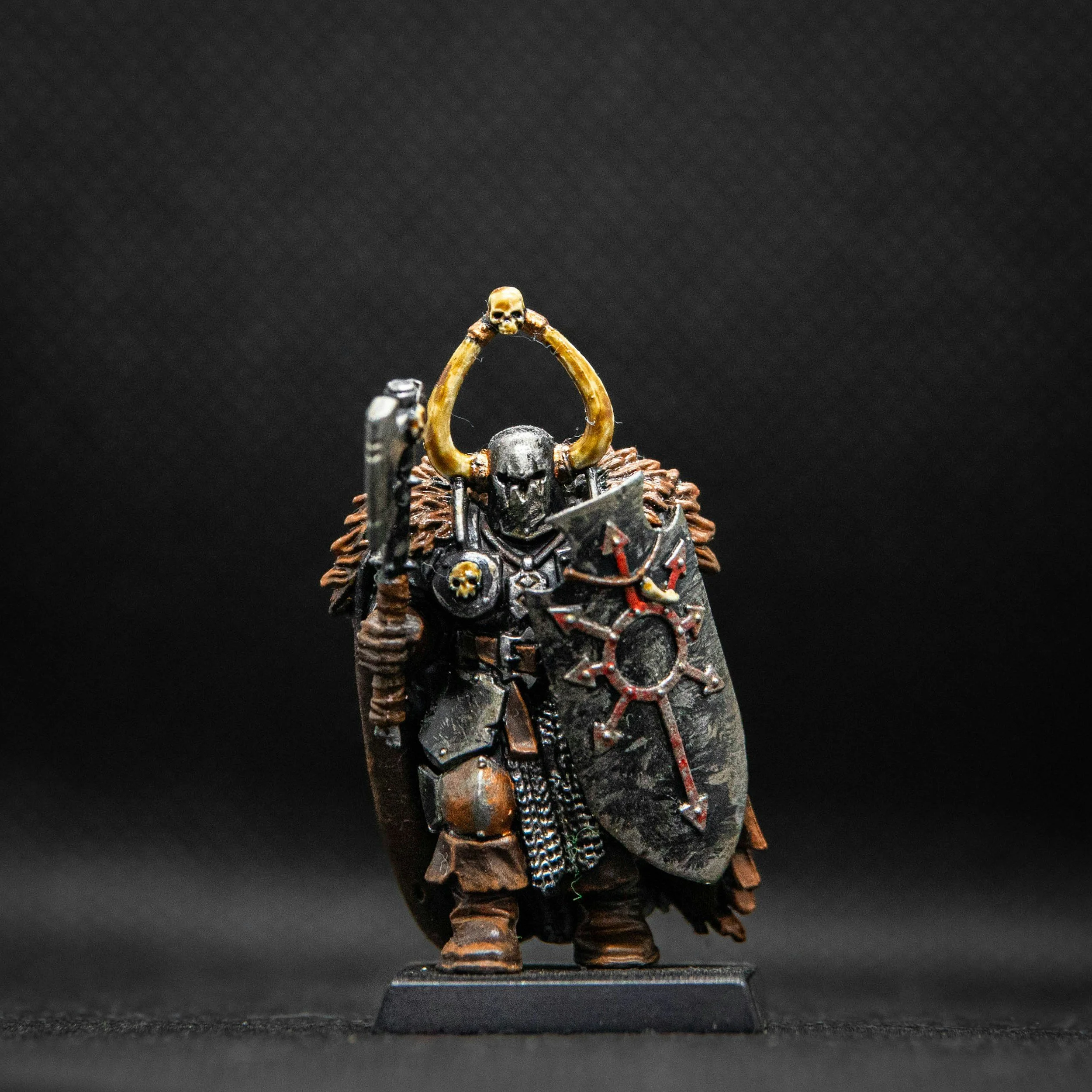 Miniature figure of a dark armored warrior with a skull helmet, holding a shield and a weapon, adorned with horns and skull accents, standing on a black base.
