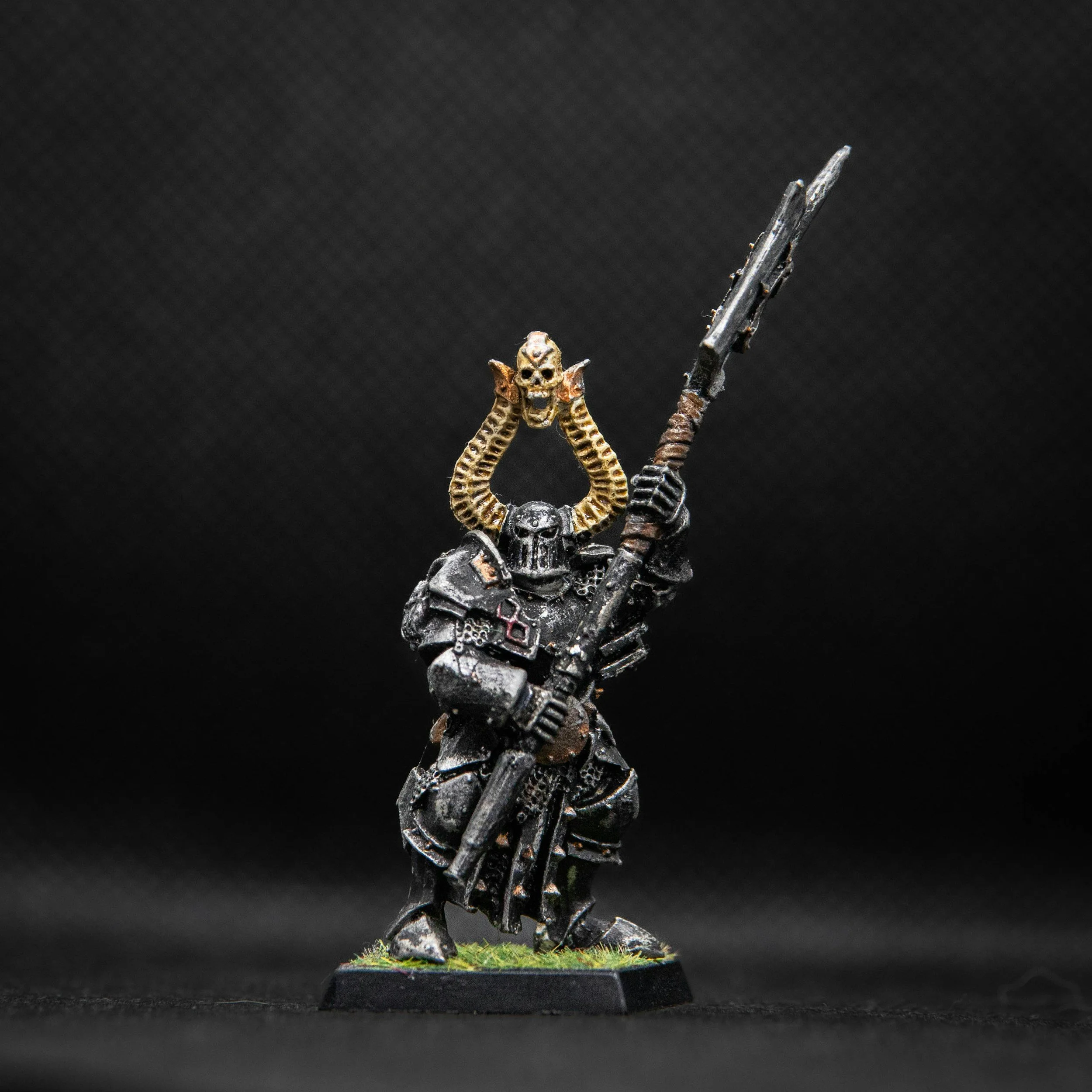 Miniature figurine of a warrior in black armor with a large spear, wearing a skull helmet with gold horns and a skull on top, standing on a grassy base.