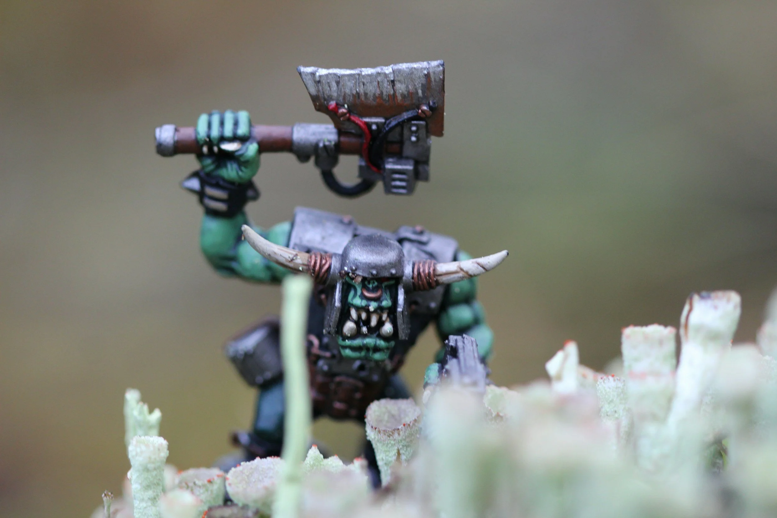 A miniature figurine of a fantasy orc warrior wielding an axe, wearing armor with horns and standing on mossy terrain.