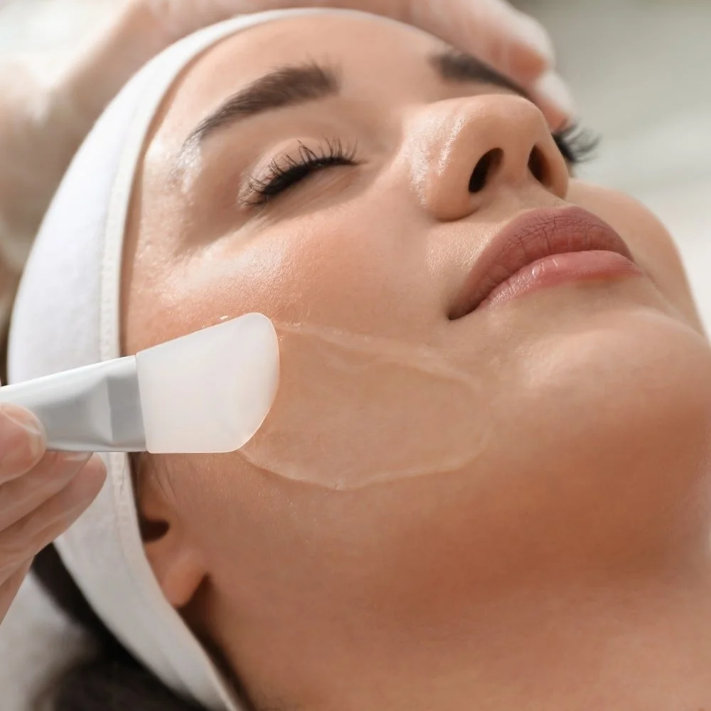 A woman receiving a facial treatment, with a skincare mask being applied to her face by a practitioner.