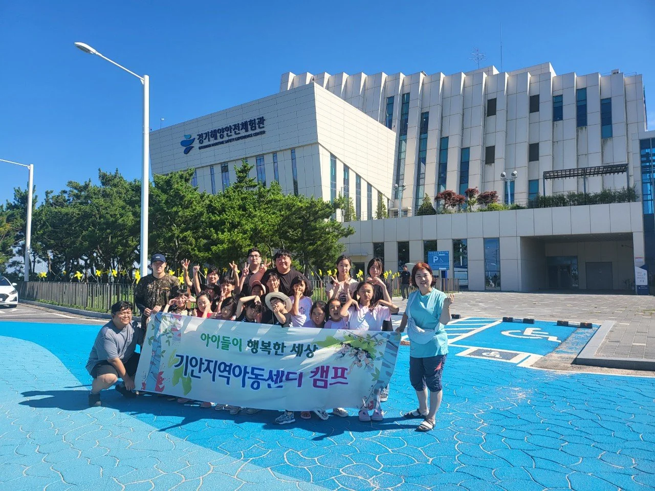 경기 아이사체 체험관 앞에서 아이들과 함께 기획지역아동센터 캠프 참여자들이 기념 촬영하는 모습, 배너에 '아이들이 행복한 세상 기획지역아동센터 캠프'라고 쓰여 있음.