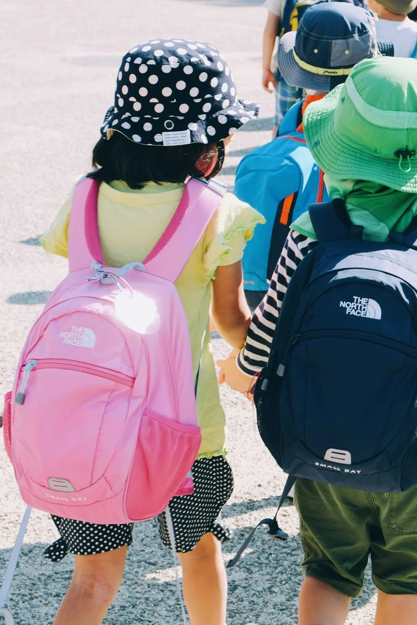 여러 어린 아이들이 야외에서 걷고 있으며, 각자 색깔이 다양한 배낭을 메고 있습니다.