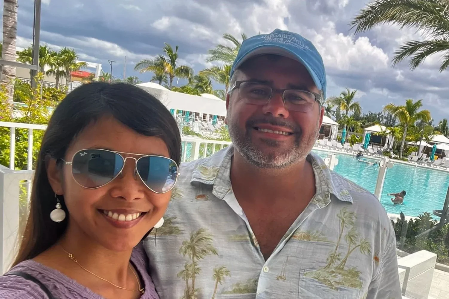 A woman and man smiling at a poolside with palm trees, lounge chairs, umbrellas, and cloudy sky in the background.