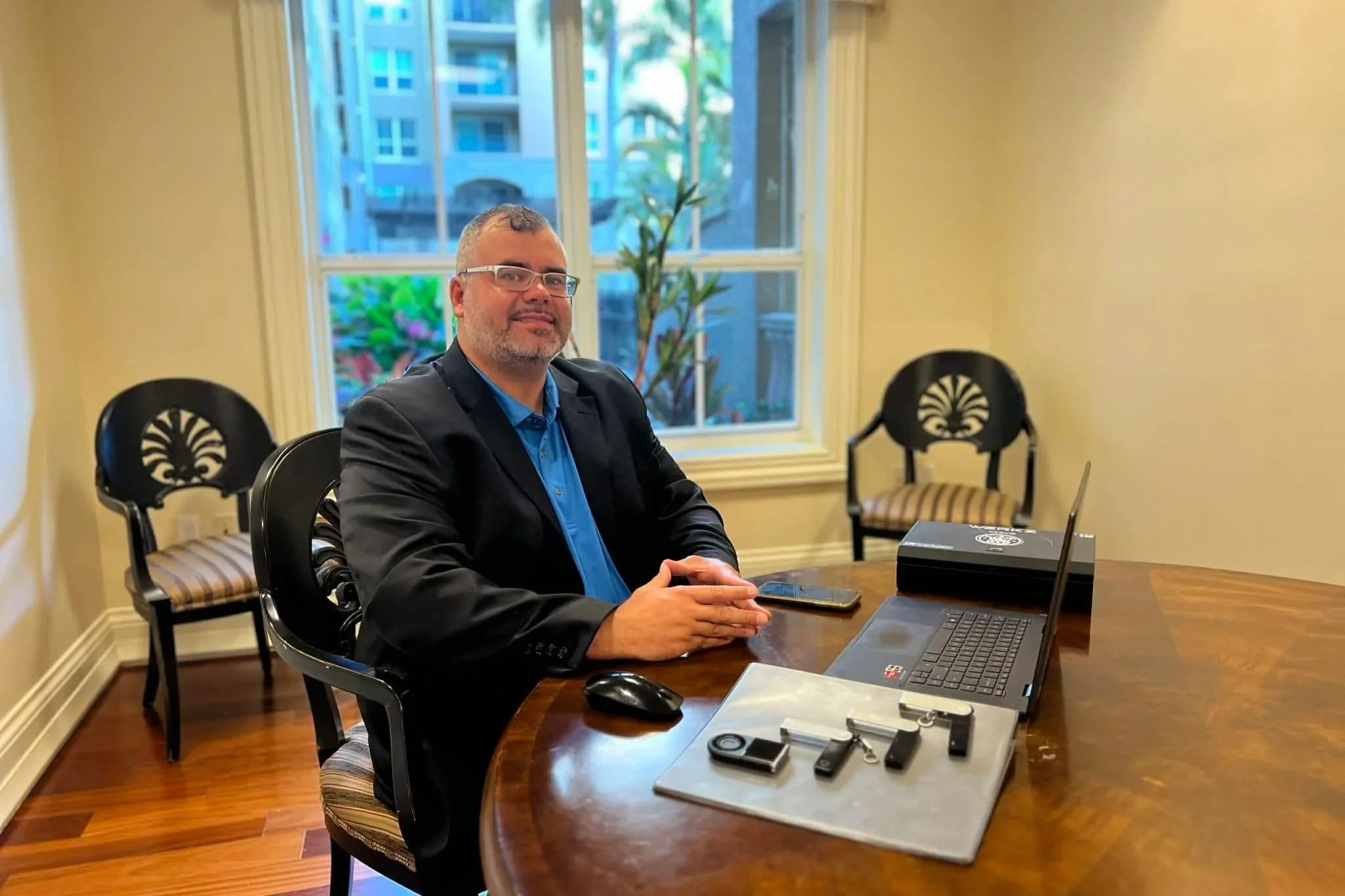 A man in a black blazer and blue shirt sitting at a wooden table with a laptop, smartphone, and various small items on it inside a room with cream yellow walls and large windows. Two black chairs with decorative backs are visible in the background.