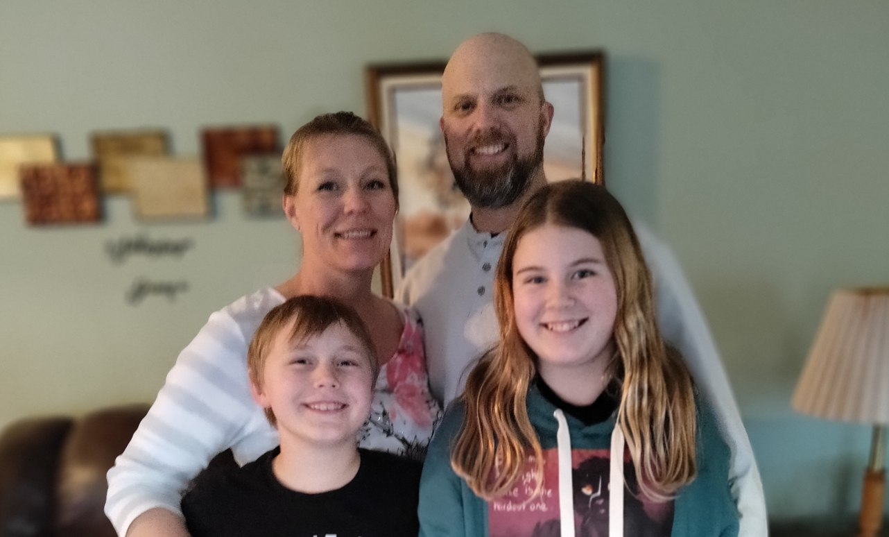 A family of four posing together indoors. The family includes a woman, a man, a young boy, and a teenage girl, all smiling at the camera with a living room background.