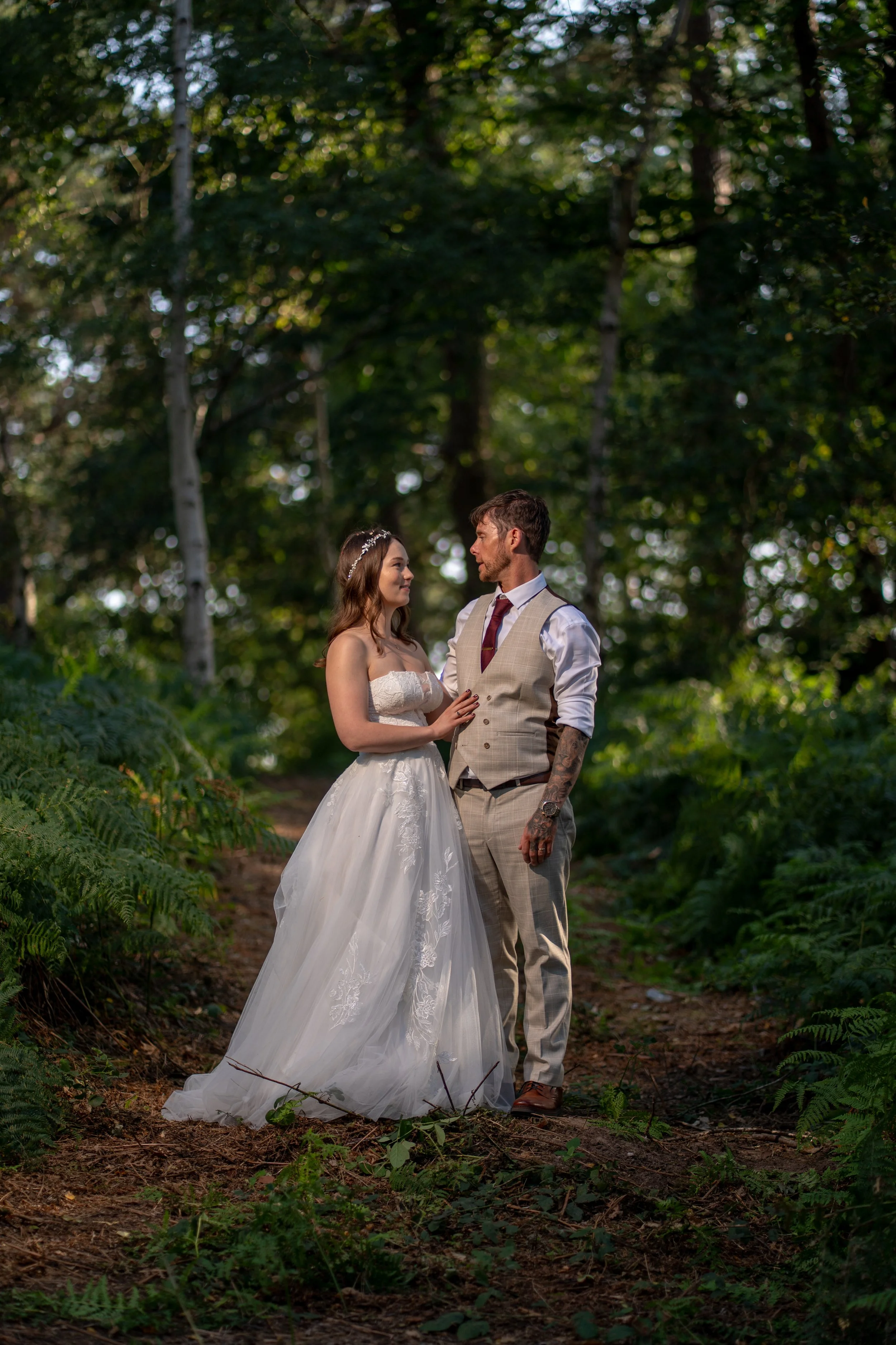 A bride and groom standing on a forest trail, gazing into each other's eyes, surrounded by green trees and ferns.