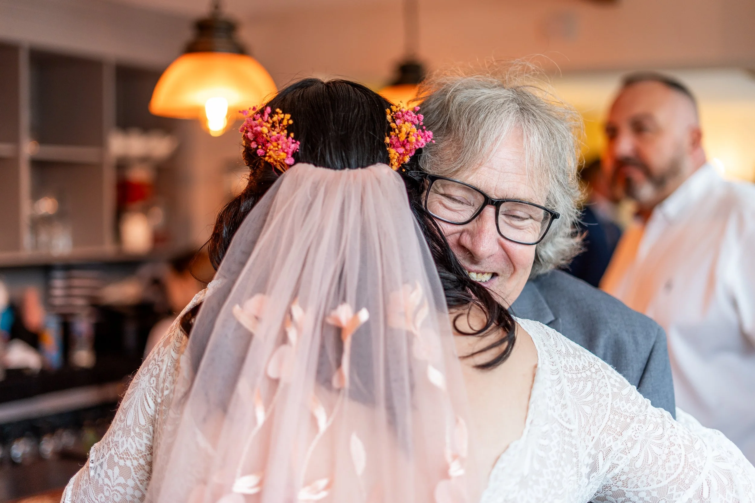 A woman in a wedding dress hugging a smiling man with glasses, surrounded by guests in a warmly lit room.