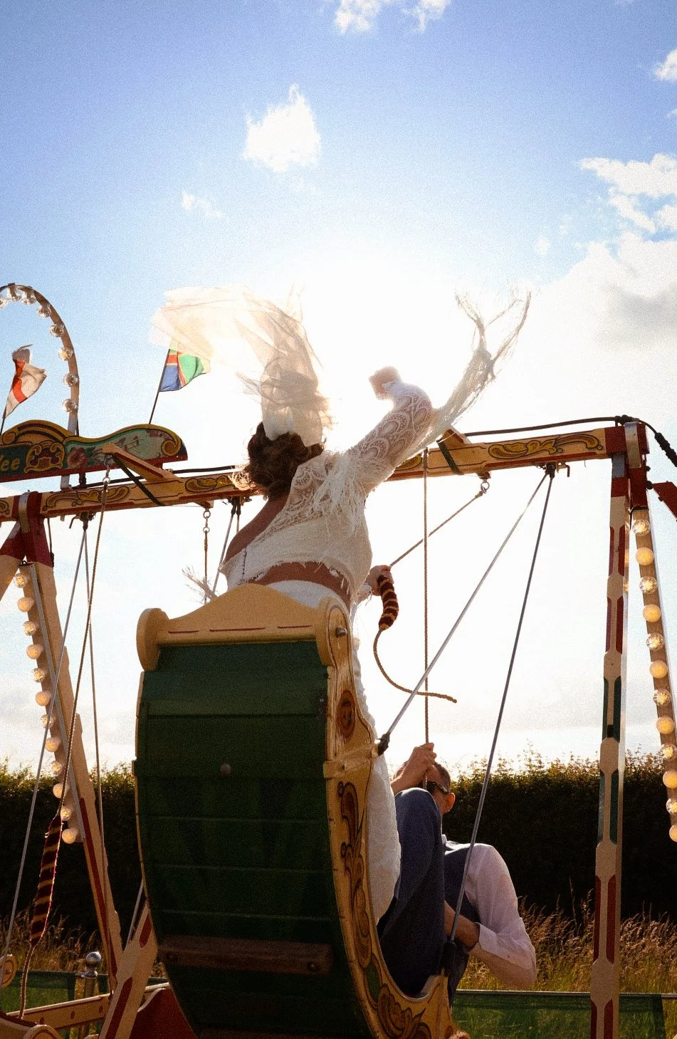 Woman in white lace dress riding a swing ride at a carnival with flags and bright sky in the background.