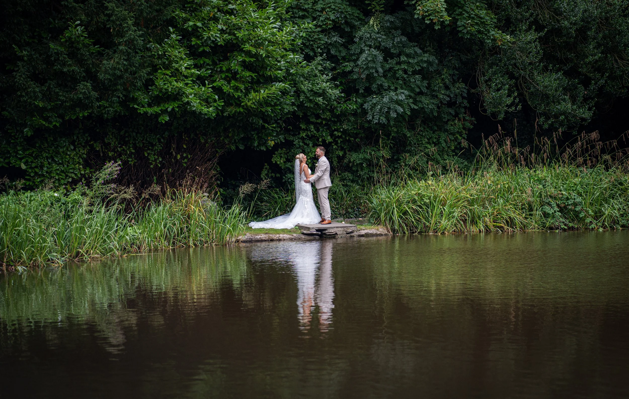 A bride and groom stand facing each other holding hands on a small stone platform by a pond, surrounded by greenery, with reflections in the water.