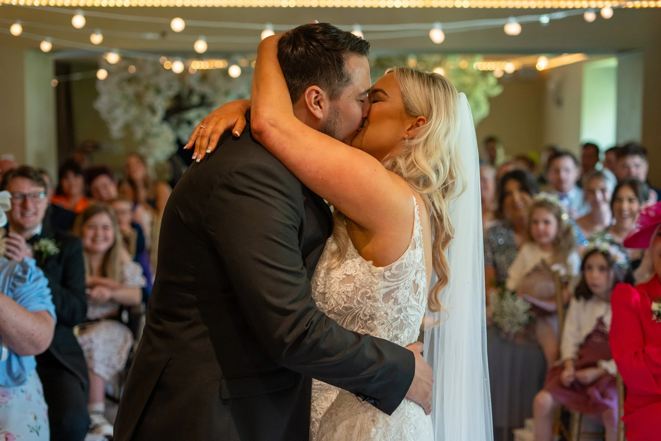 A bride and groom kiss during their wedding ceremony, surrounded by seated guests watching happily.