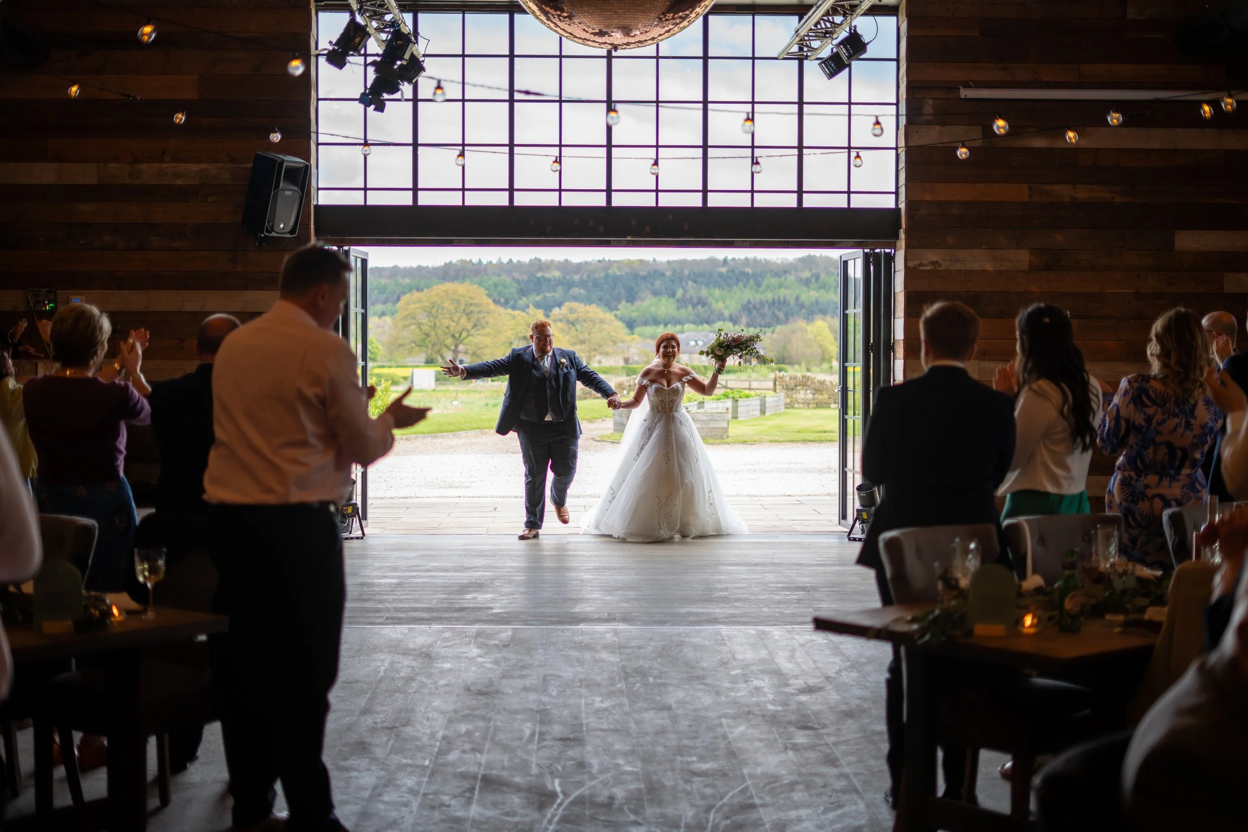 Bride and groom entering a rustic barn wedding venue holding hands and smiling, with guests clapping and celebrating inside.