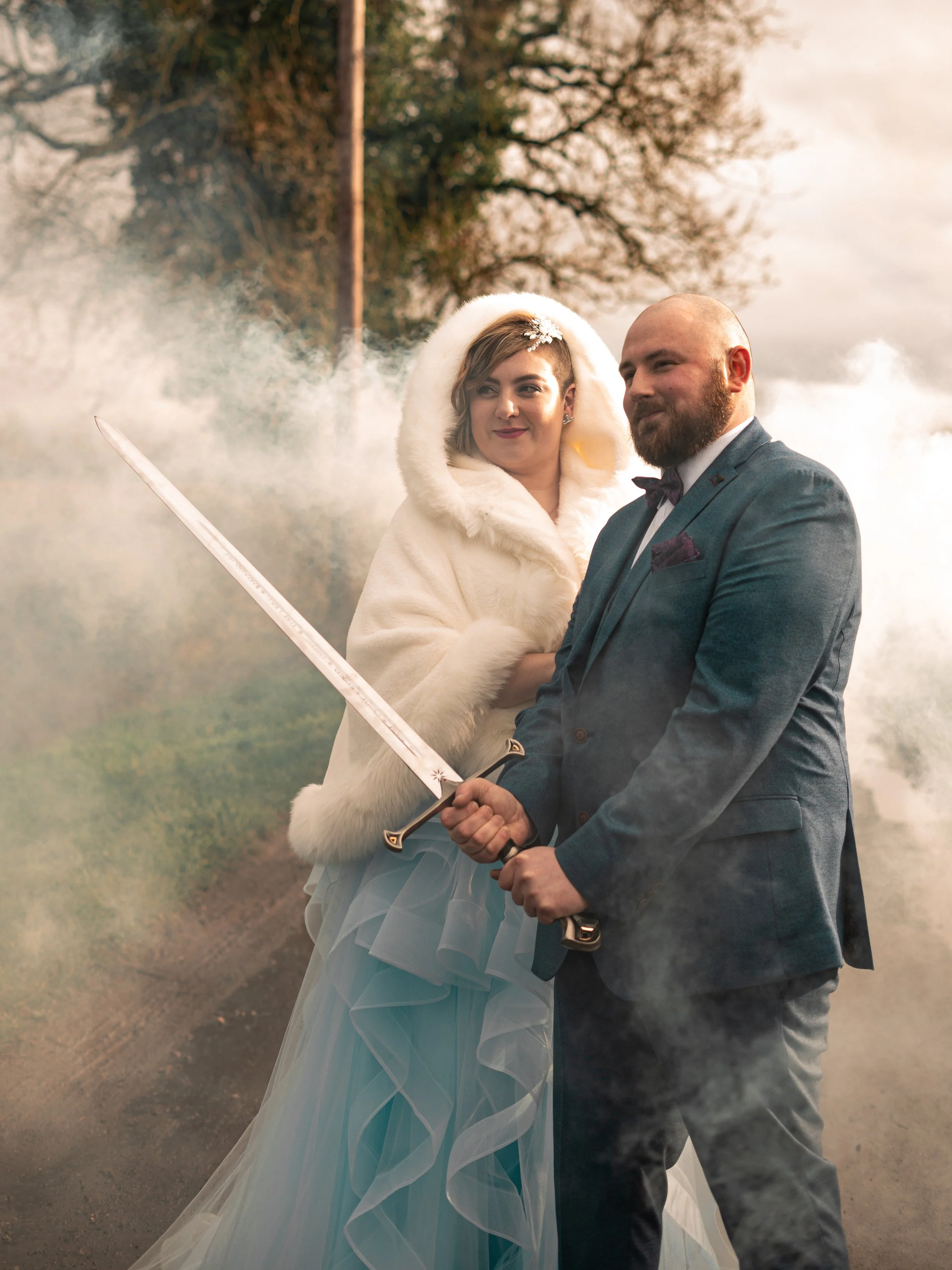 A bride and groom dressed in wedding attire, holding a sword with a misty outdoor setting and trees in the background.