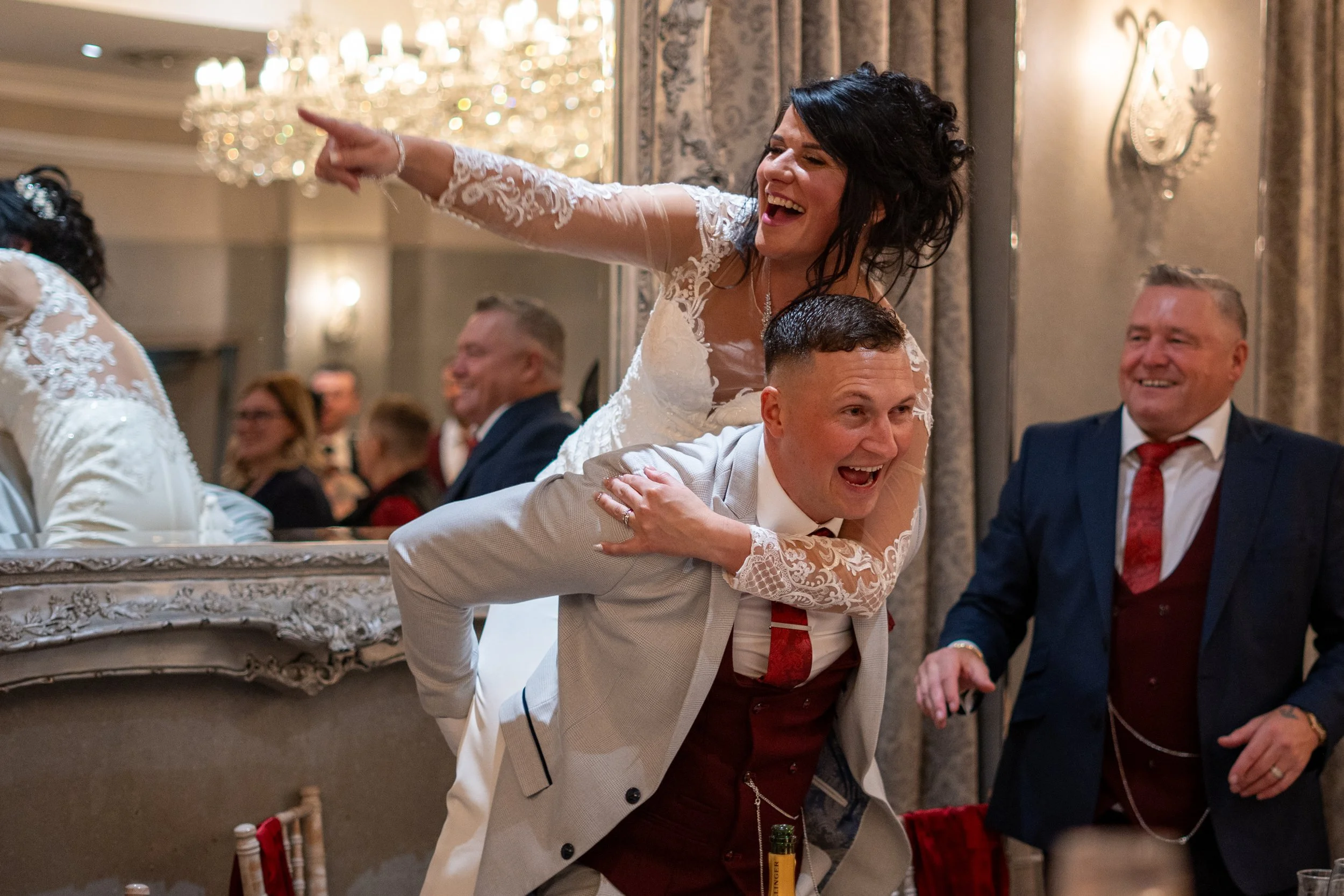 People at a wedding celebration, with a woman in a wedding dress piggyback riding a man, both smiling and laughing. Others in formal attire are visible in the background.