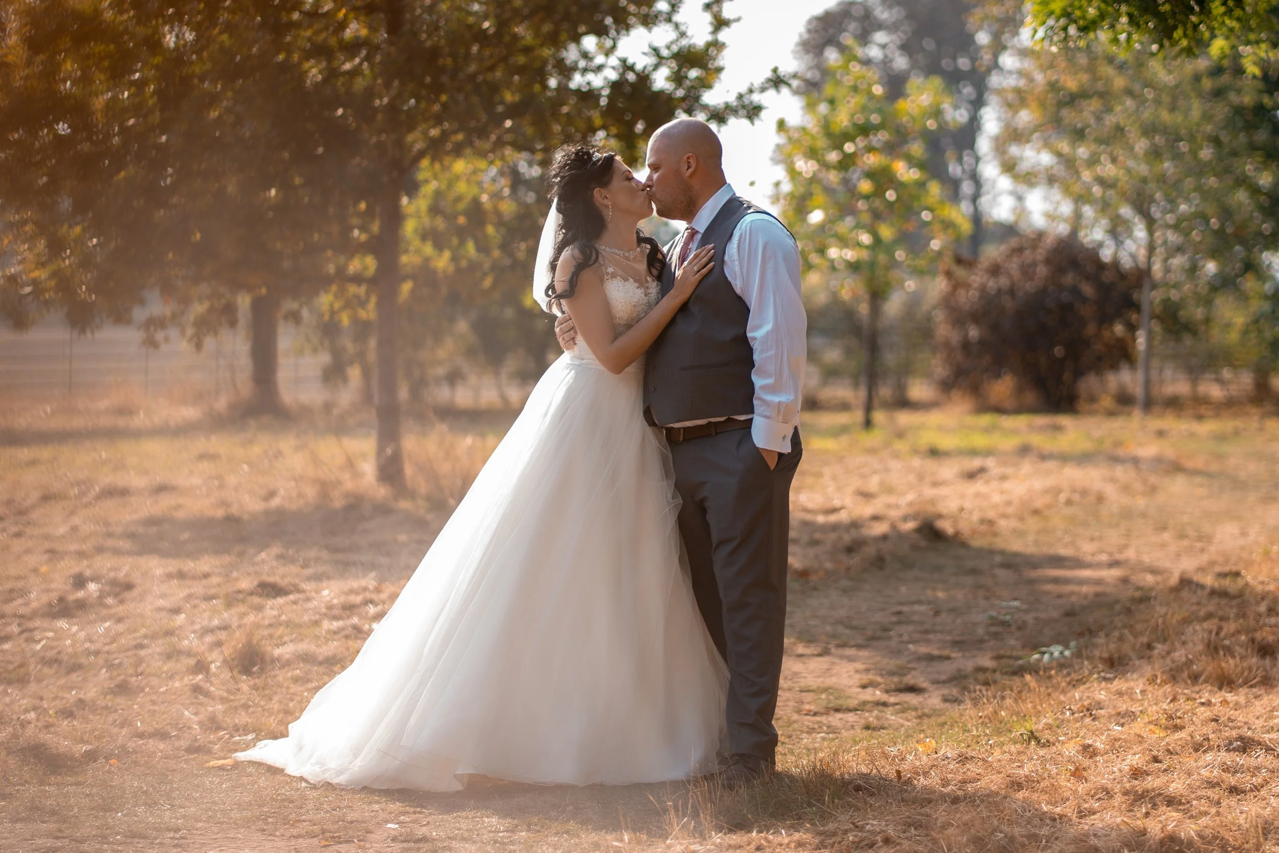 A bride and groom kissing outdoors in a wooded area during sunset, with trees in the background and fallen leaves on the ground.
