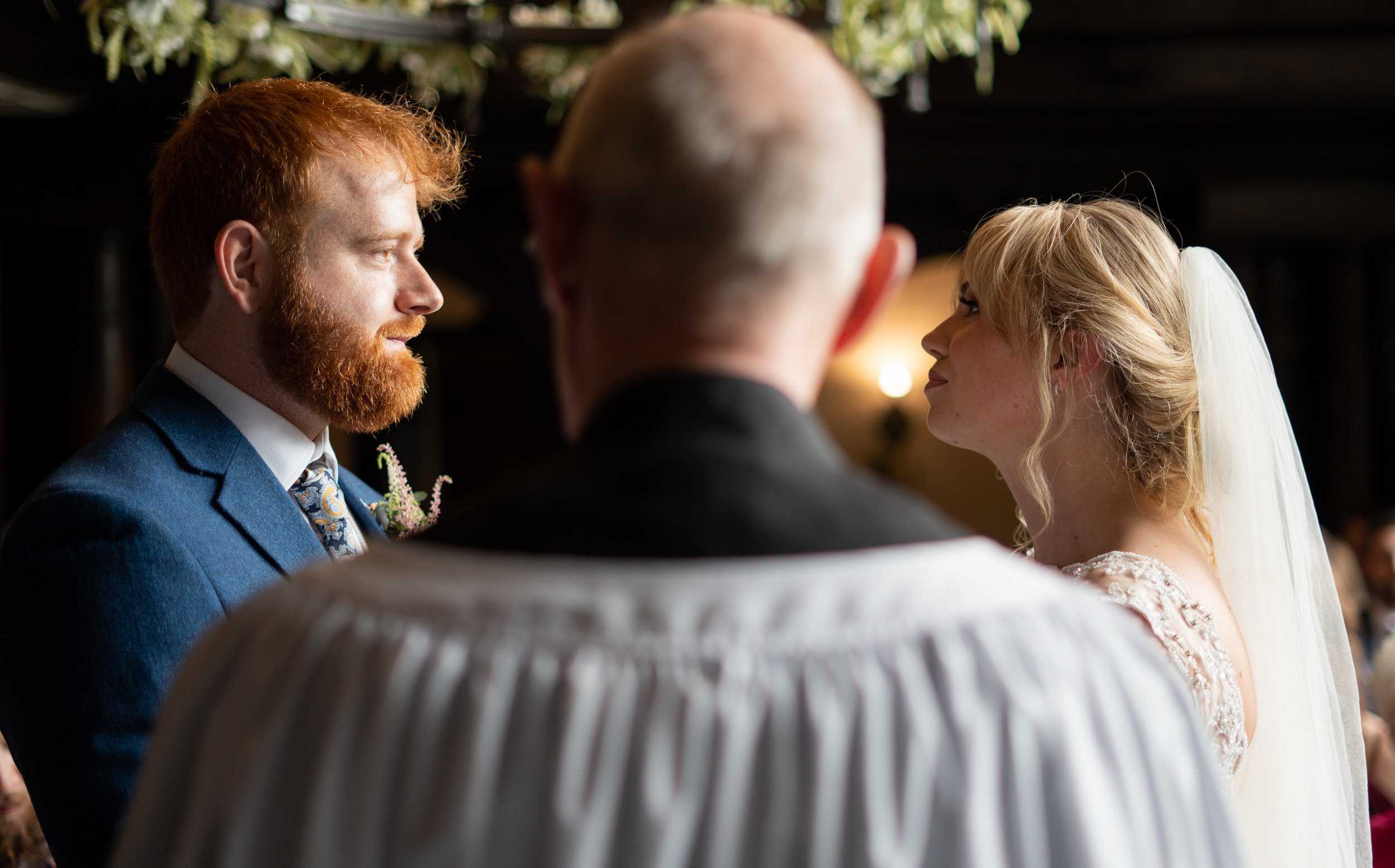 Bride and groom facing each other during their wedding ceremony, with officiant in foreground, in a dimly lit indoor setting.