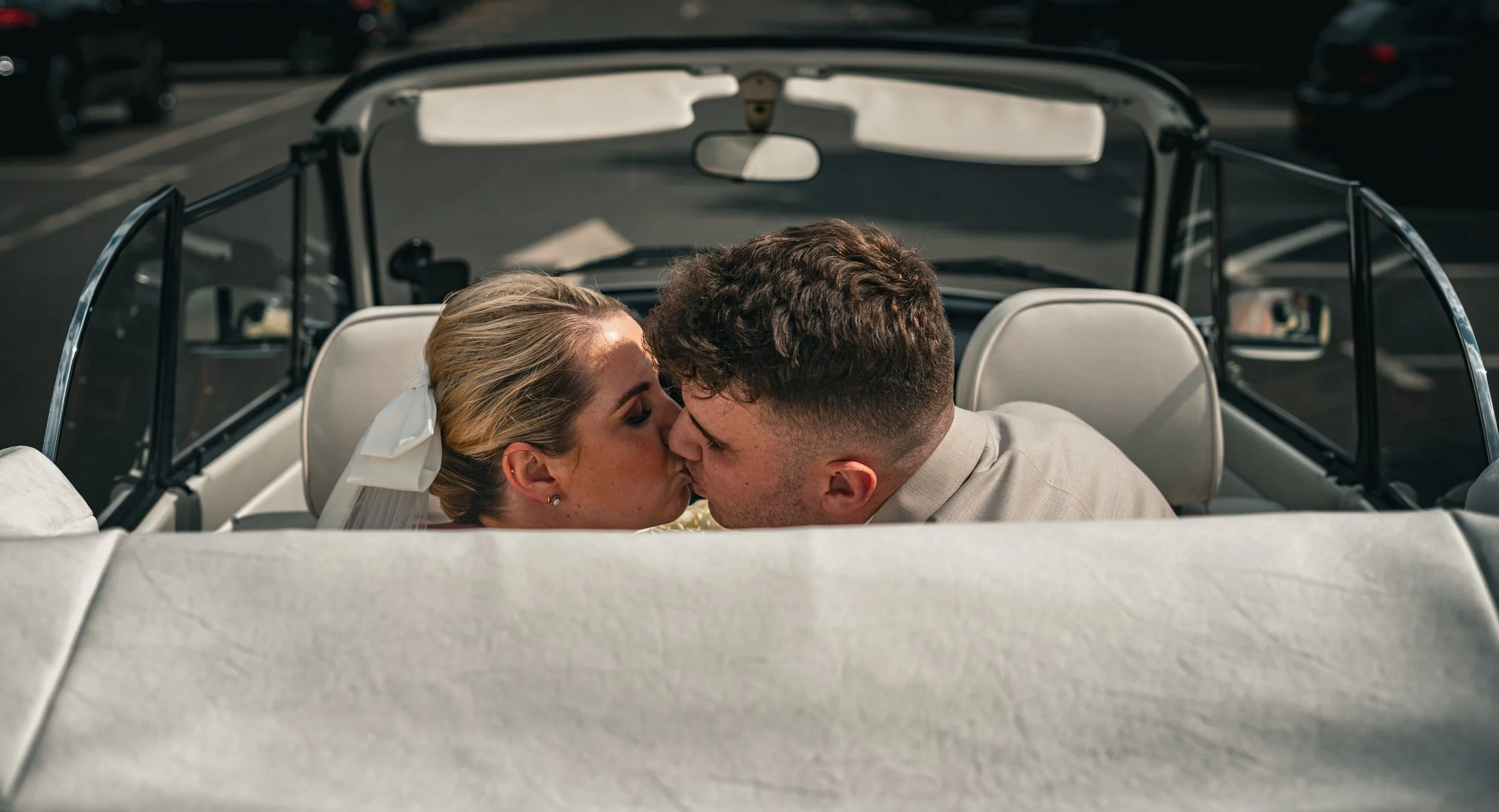 A newlywed couple in wedding attire sharing a kiss in the back of a vintage convertible car.