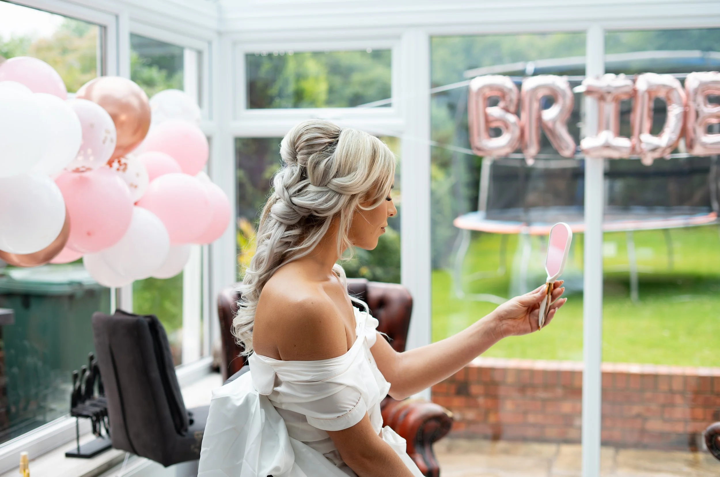 A woman with long, curly, platinum blonde hair and wearing a white off-shoulder dress sits in a sunroom with large windows, holding a pink handheld mirror, with balloons in pink, white, and gold and a 'BRIDE' banner in the background.