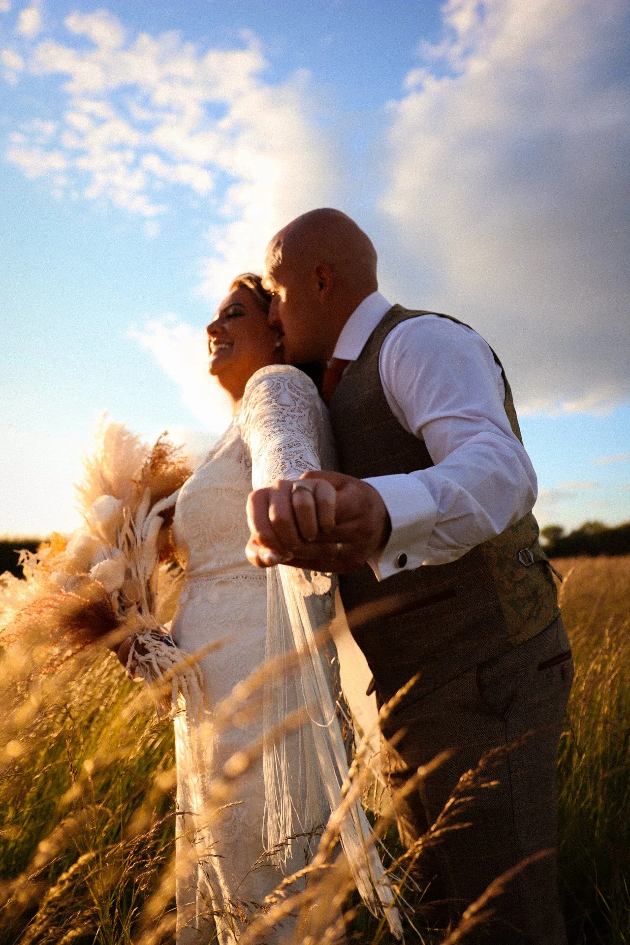 A happy couple dancing and holding hands in a field at sunset, with the sky partly cloudy in the background.