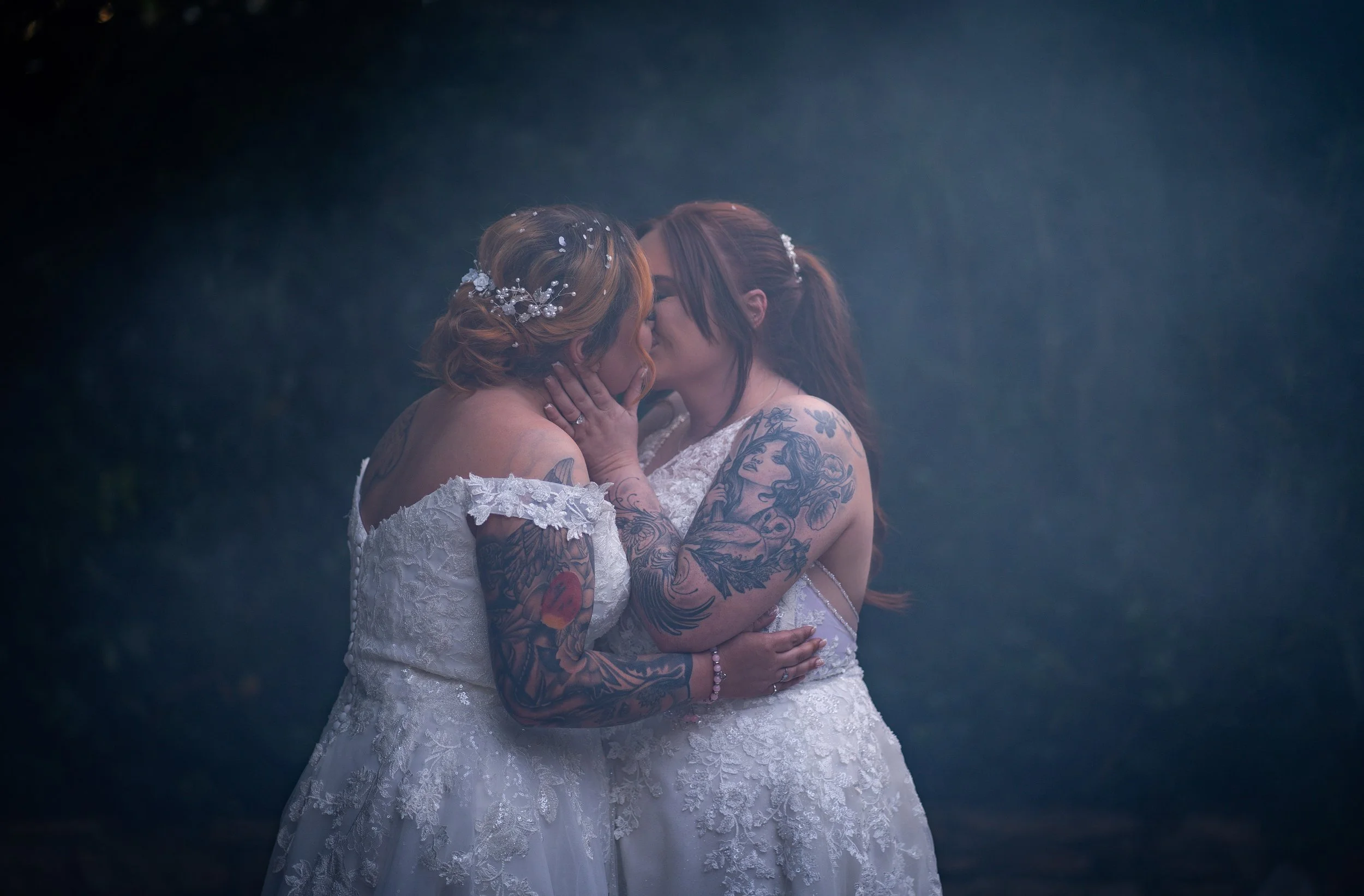 Two women in wedding dresses kissing each other outdoors, surrounded by fog, with their tattoos visible.