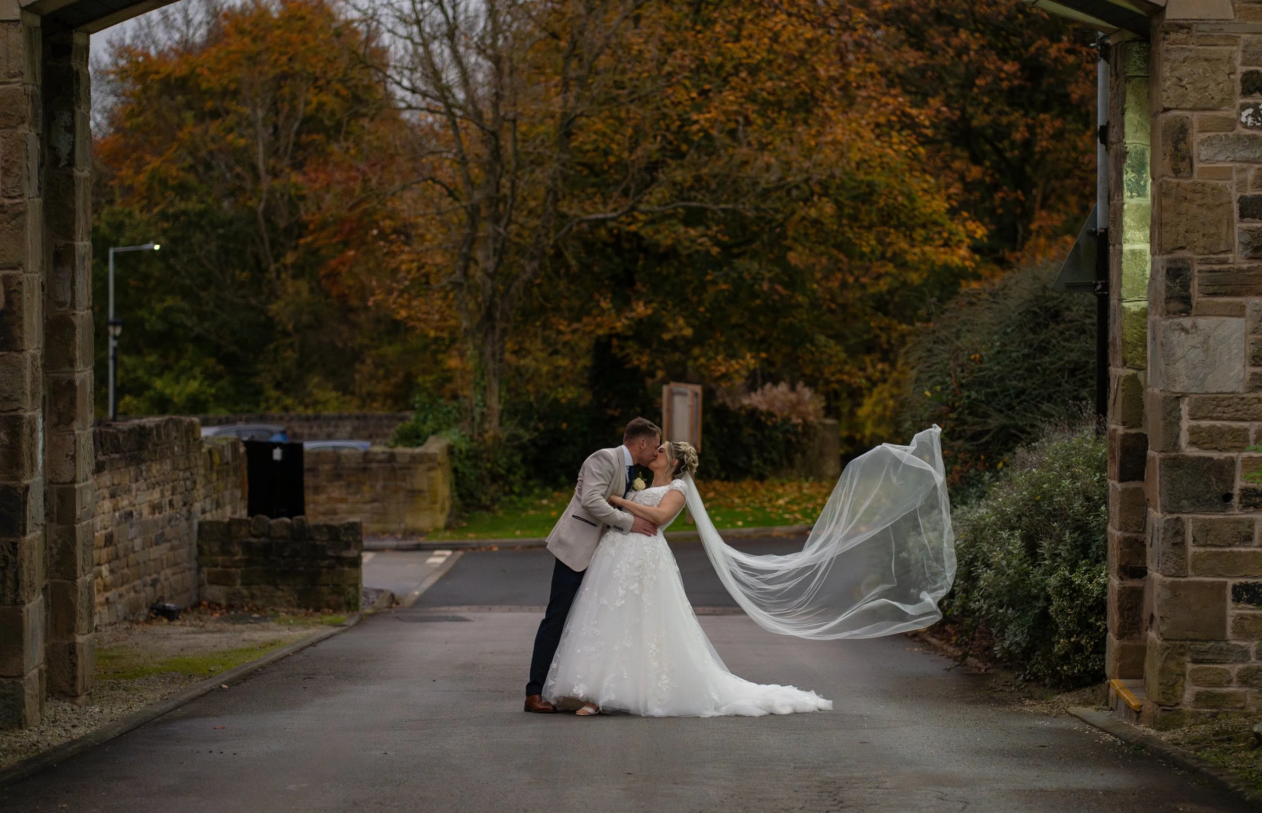 A bride and groom sharing a kiss under an archway on their wedding day, with autumn trees in the background.