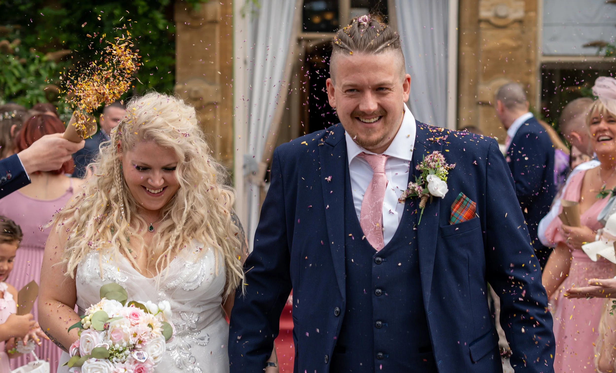 A happy bride and groom walking outdoors at their wedding reception, surrounded by guests throwing confetti, with a rustic building and curtains in the background.