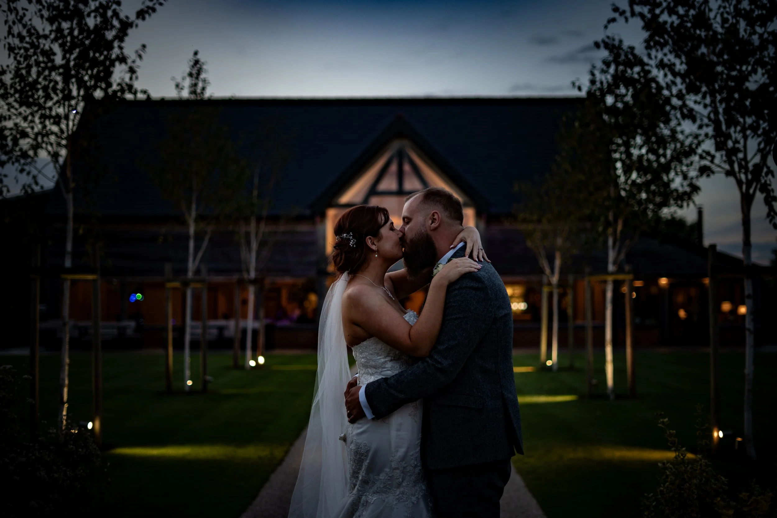 A bride and groom sharing a kiss outdoors during sunset, with a barn-style building in the background.