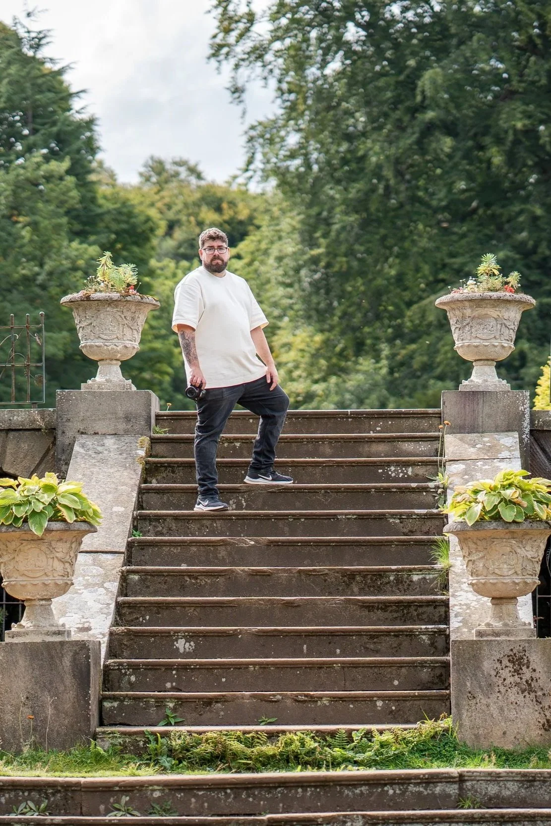 A man with glasses, beard, and tattoos on his left arm standing on outdoor stone stairs with large planter flower pots on either side, surrounded by greenery and trees.