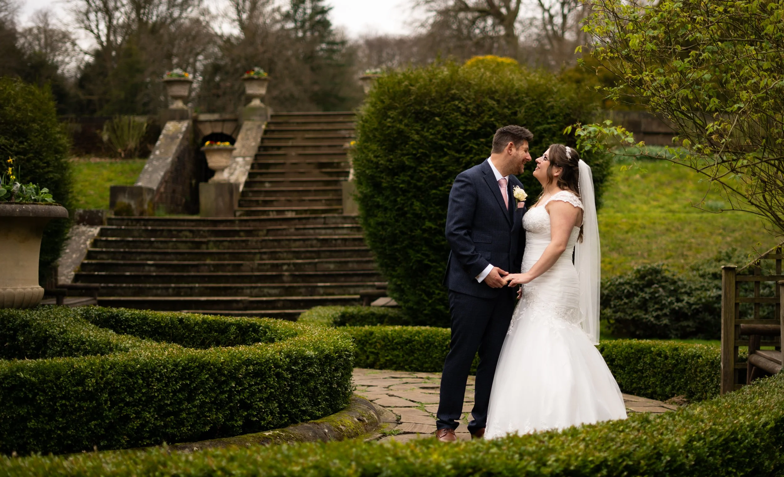 A bride and groom holding hands, smiling at each other outdoors in a garden setting.